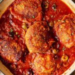 Chicken Puttanesca thighs cooked in a rich tomato sauce with olives and herbs in a white pot, surrounded by bread and seasonings.