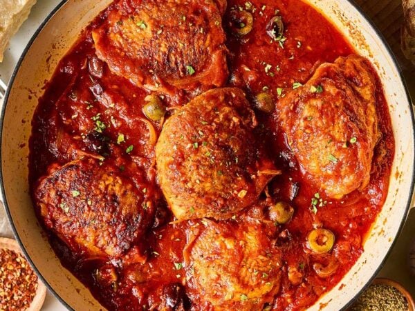 Chicken Puttanesca thighs cooked in a rich tomato sauce with olives and herbs in a white pot, surrounded by bread and seasonings.