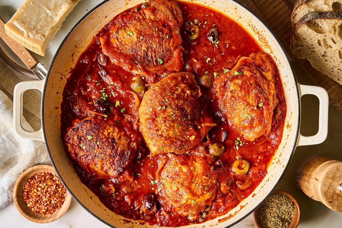 Chicken Puttanesca thighs simmer in tomato sauce with olives and herbs in a white Dutch oven, surrounded by bread and spices.