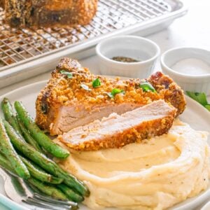 Homemade Shake and Bake Pork Chops served atop mashed potatoes with green beans, accompanied by salt, pepper, and parsley on the side.