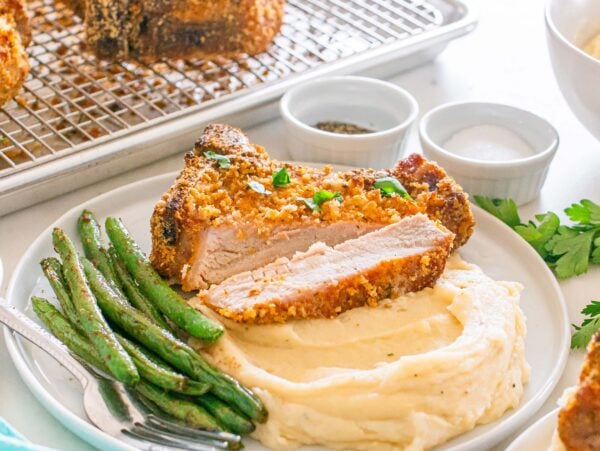 Homemade Shake and Bake Pork Chops served atop mashed potatoes with green beans, accompanied by salt, pepper, and parsley on the side.