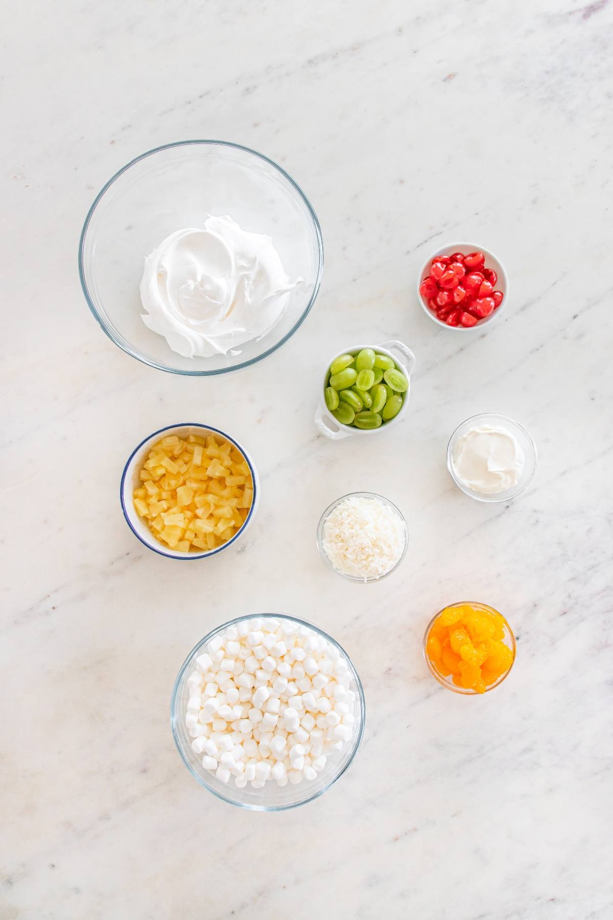 Eight bowls with ingredients: whipped topping, cherries, pineapple, marshmallows, coconut, oranges, nuts, sour cream.