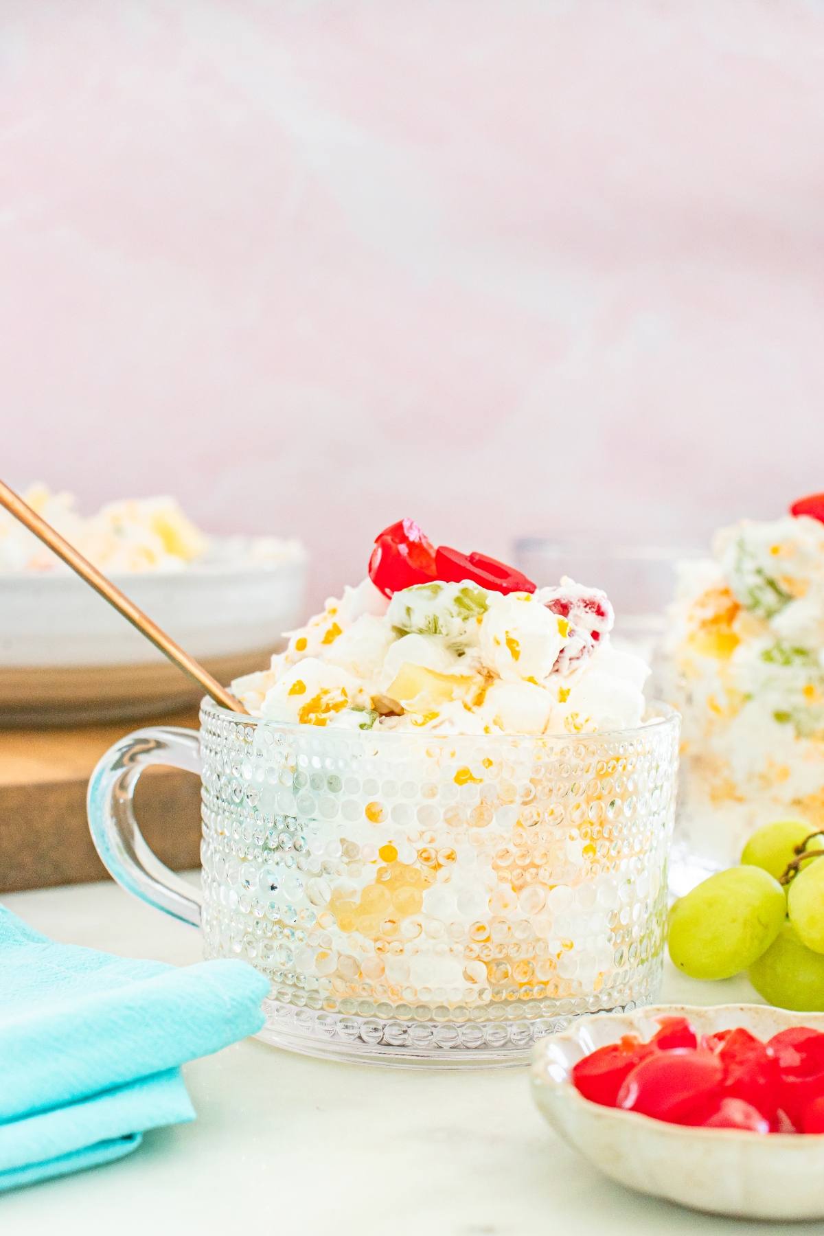 A glass cup of creamy ambrosia fruit salad topped with cherries, with grapes and chopped fruit nearby on a table.