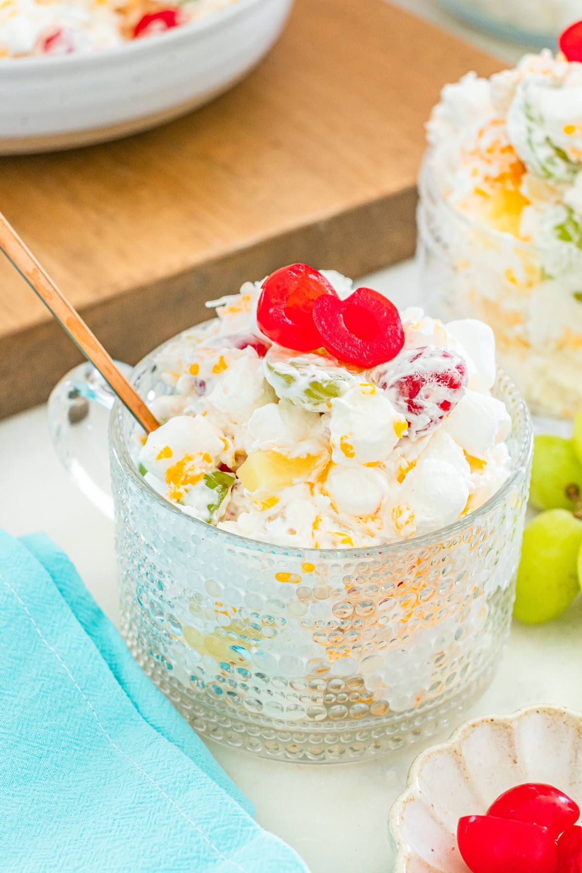 A glass bowl of ambrosia fruit salad with marshmallows, grapes, pineapple, and cherries, with a spoon inside.