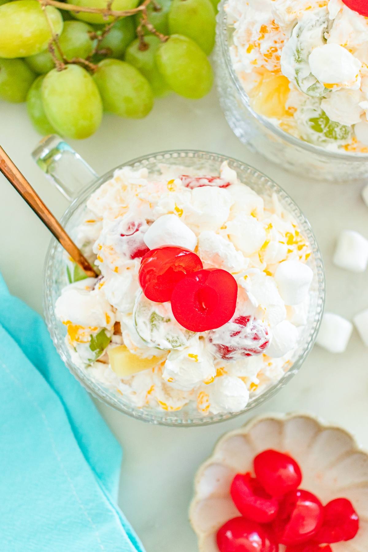 A glass bowl of ambrosia fruit salad topped with cherries, with grapes and marshmallows nearby.