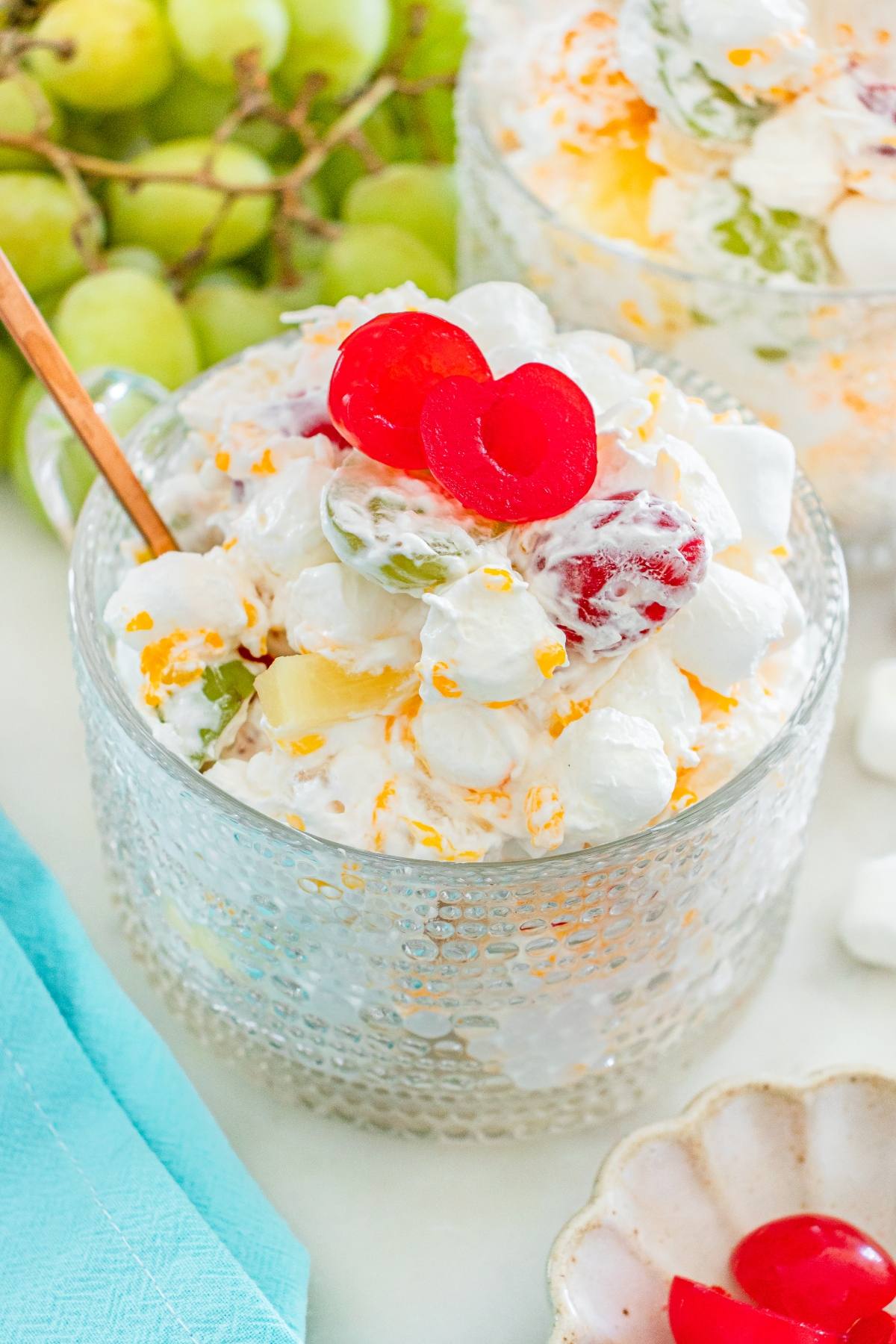 A glass bowl of ambrosia fruit salad with marshmallows, grapes, pineapple, and cherries, topped with two cherry halves.