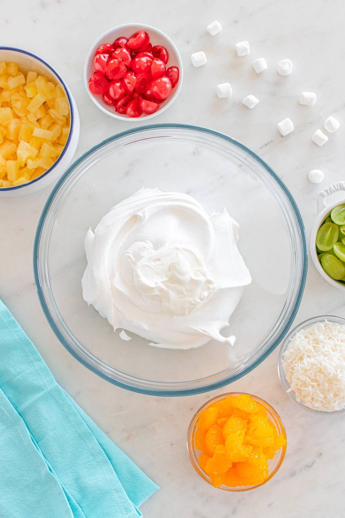A bowl of whipped topping surrounded by bowls of pineapple, cherries, oranges, limes, coconut, and mini marshmallows.