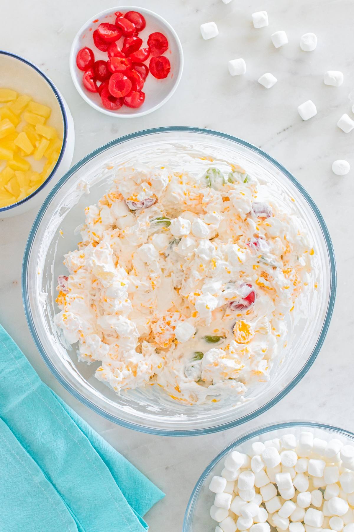 Large glass bowl with fruit salad, surrounded by cherries, pineapple, marshmallows, and a blue napkin.