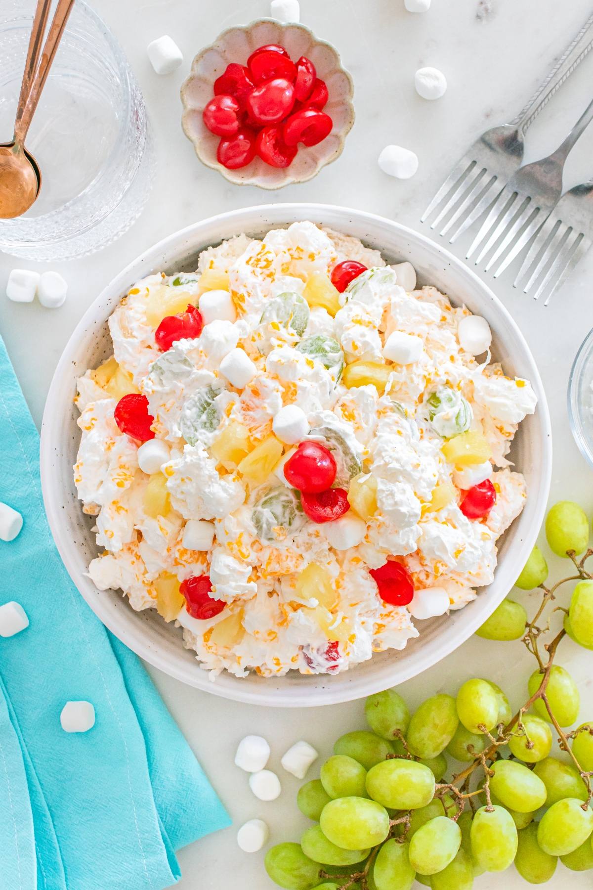 A bowl of creamy ambrosia fruit salad with cherries, pineapple, grapes, and mini marshmallows, surrounded by green grapes.