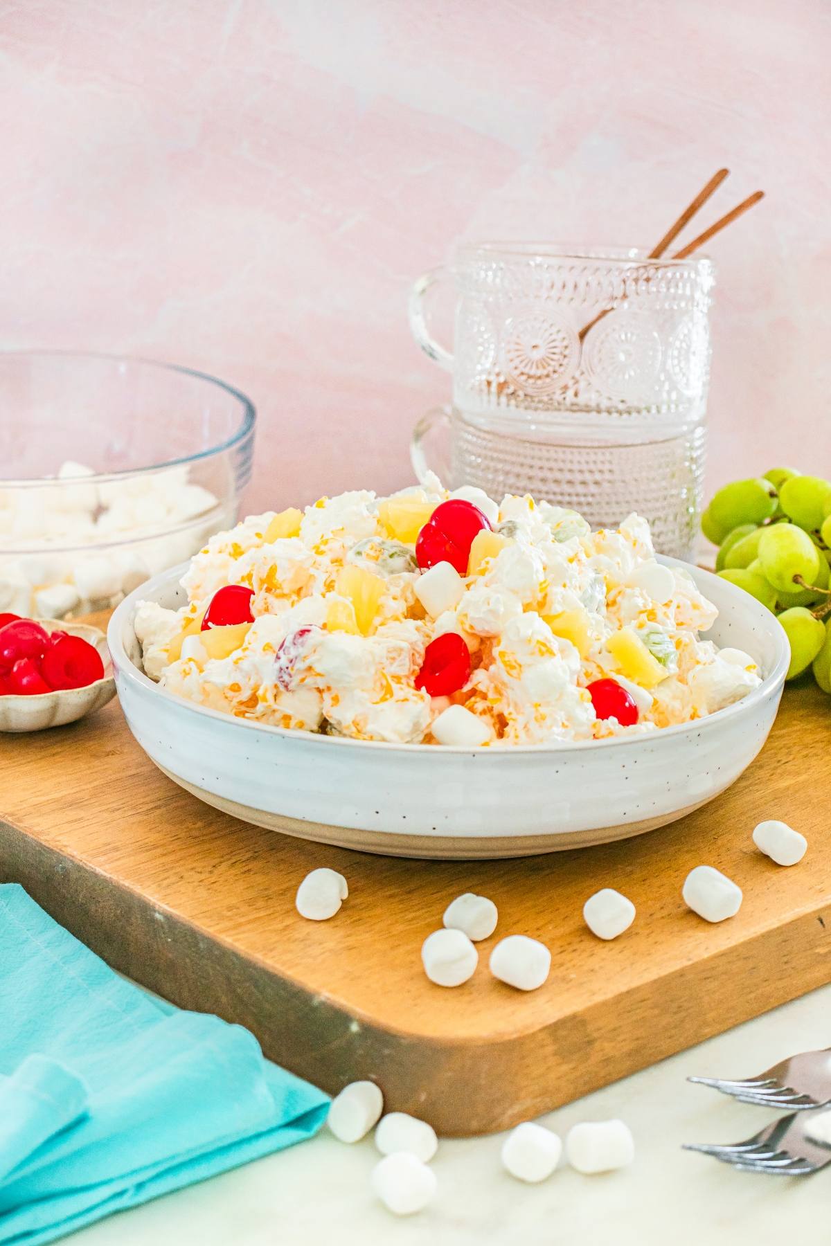A bowl of ambrosia fruit salad with marshmallows and cherries, next to grapes, cups, and serving utensils on a board.