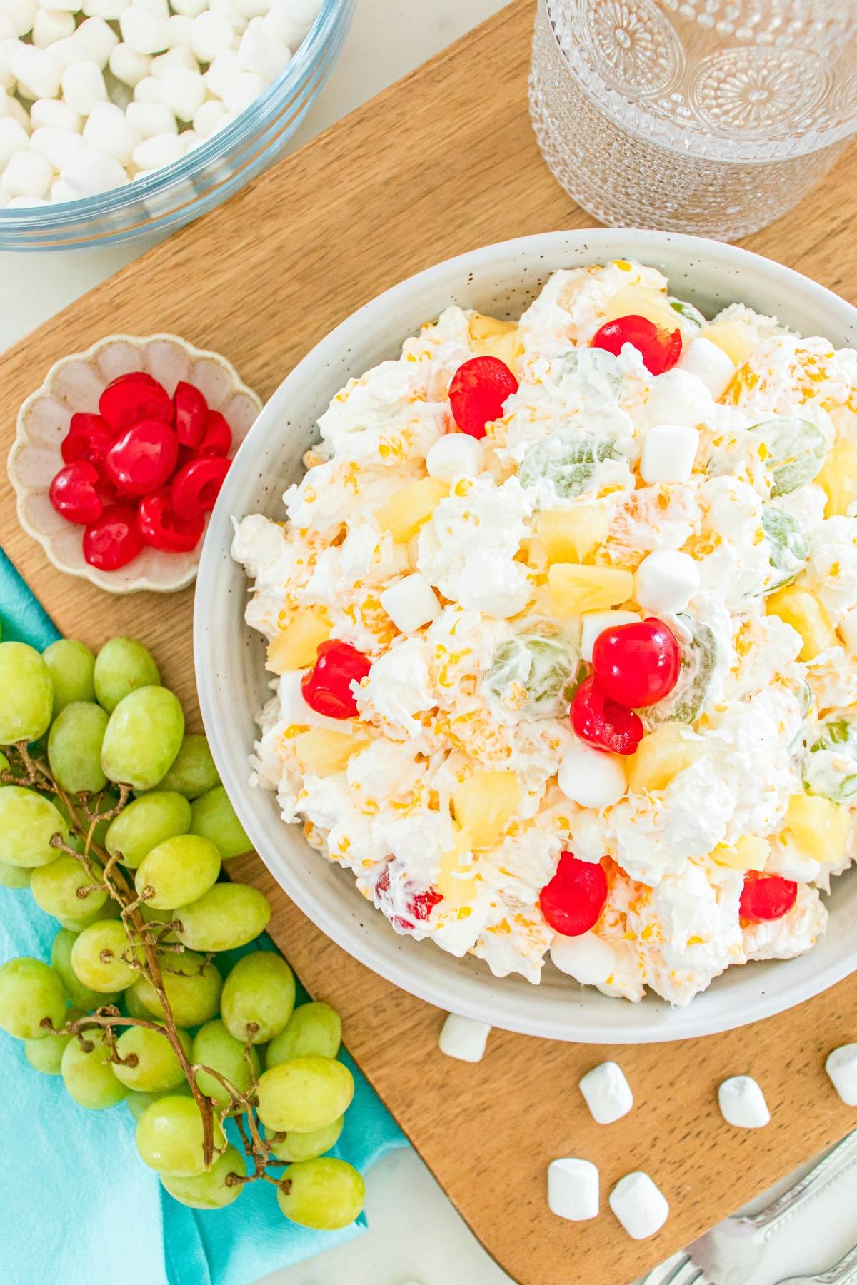 A bowl of creamy ambrosia fruit salad with cherries, marshmallows, and grapes on a wooden board beside green grapes.