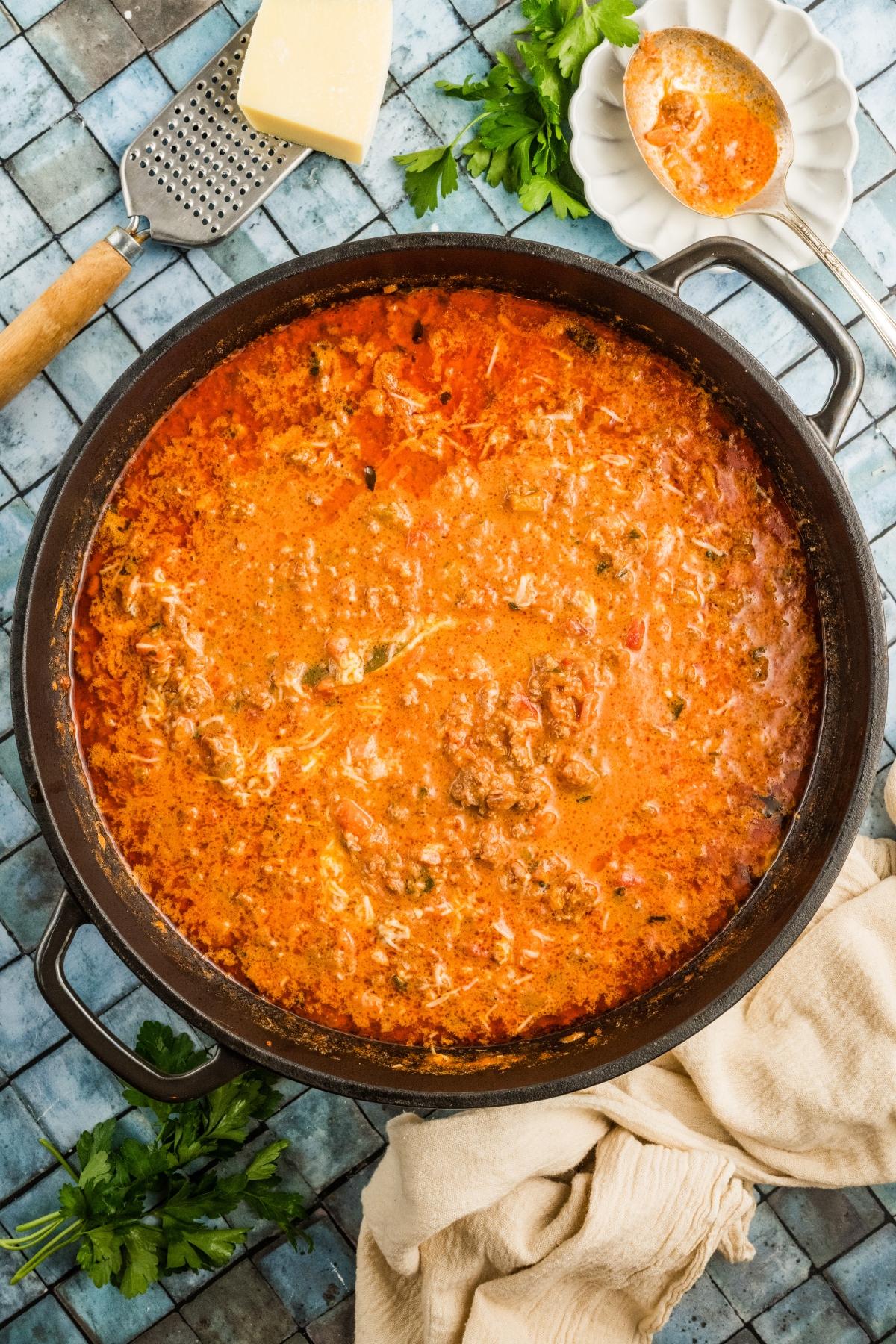 Large pot filled with creamy tomato and meat sauce, surrounded by Parmesan, parsley, and a grater on a tile surface.