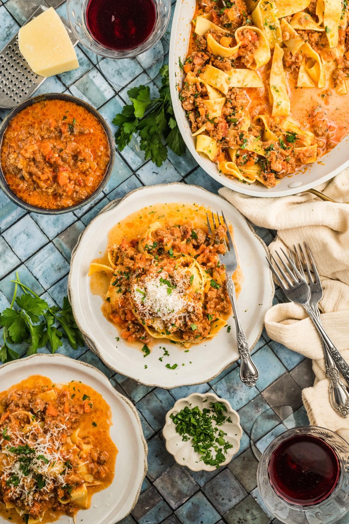 Overhead view of pasta with meat authentic bolognese sauce, grated cheese, wine glasses, and fresh herbs on a tiled table.