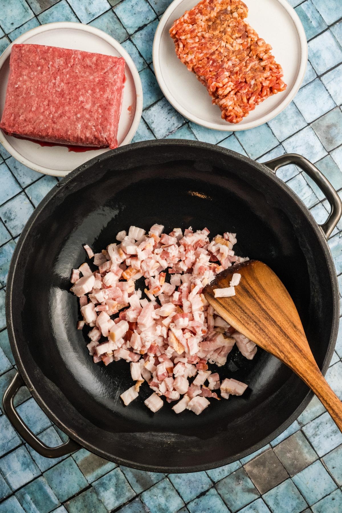 Chopped bacon cooking in a black pan, with ground beef and sausage on plates nearby.