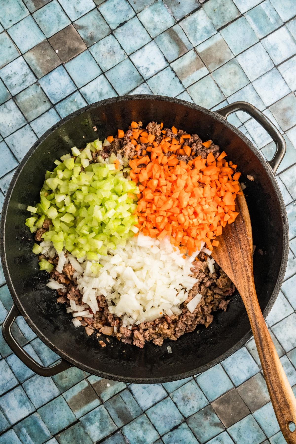 Chopped celery, carrots, and onions in a skillet with ground beef, stirred with a wooden spoon.