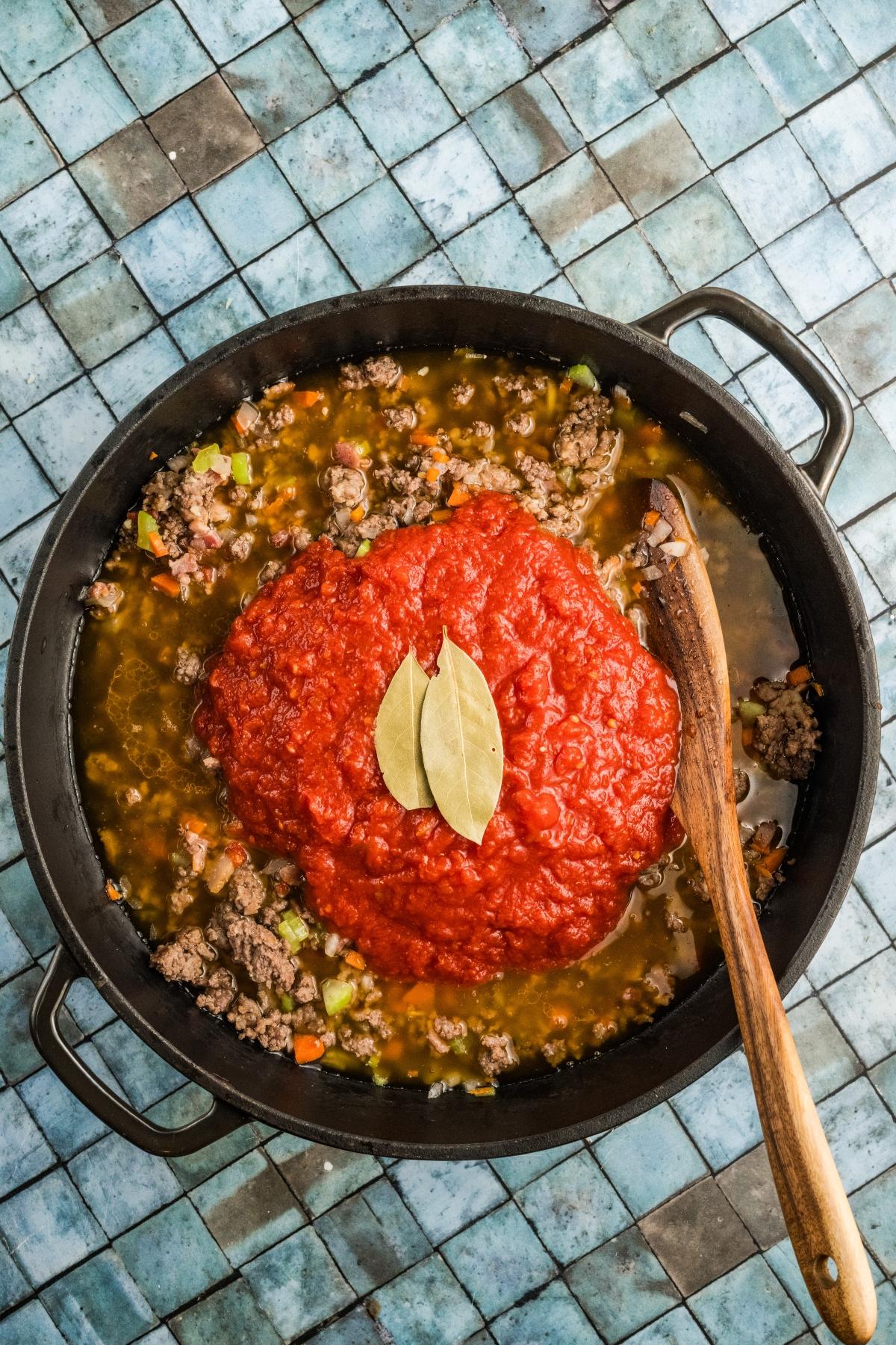 A pot of ground meat and vegetables topped with tomato sauce and two bay leaves, on a blue tiled surface.