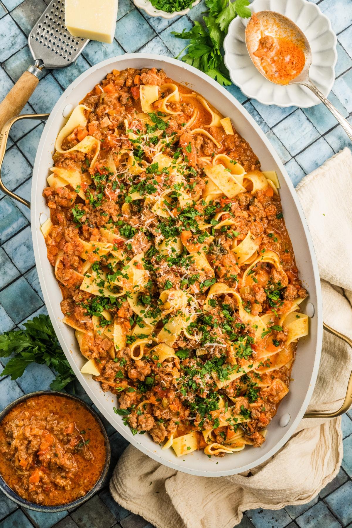 Large oval dish of pappardelle pasta with meat authentic bolognese sauce, topped with parsley and grated cheese on a blue surface.