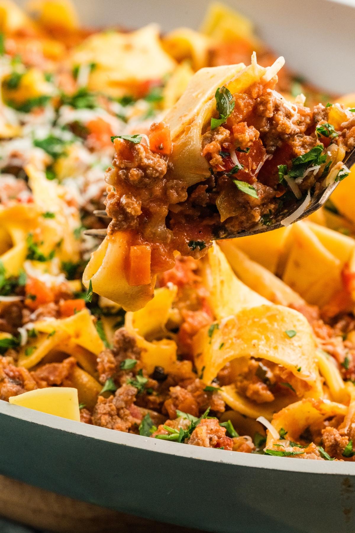 A fork lifts pappardelle pasta with meat sauce and herbs from a pan.