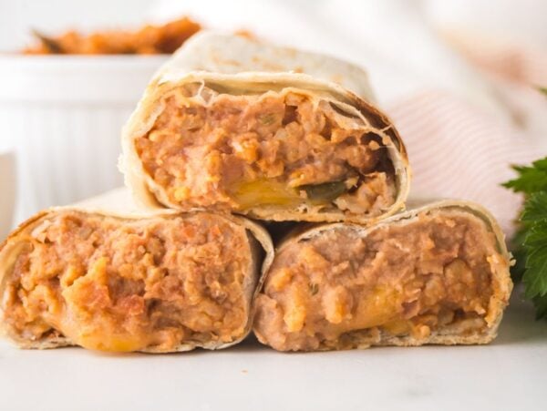 Three bean and cheese burritos stacked on a white surface with a garnish of fresh parsley nearby, highlighting the classic appeal of bean and cheese burritos.
