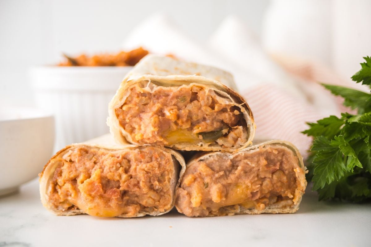 Three bean and cheese burritos are stacked on a white surface, with a bowl and fresh herbs in the background, highlighting these flavorful bean and cheese burritos.