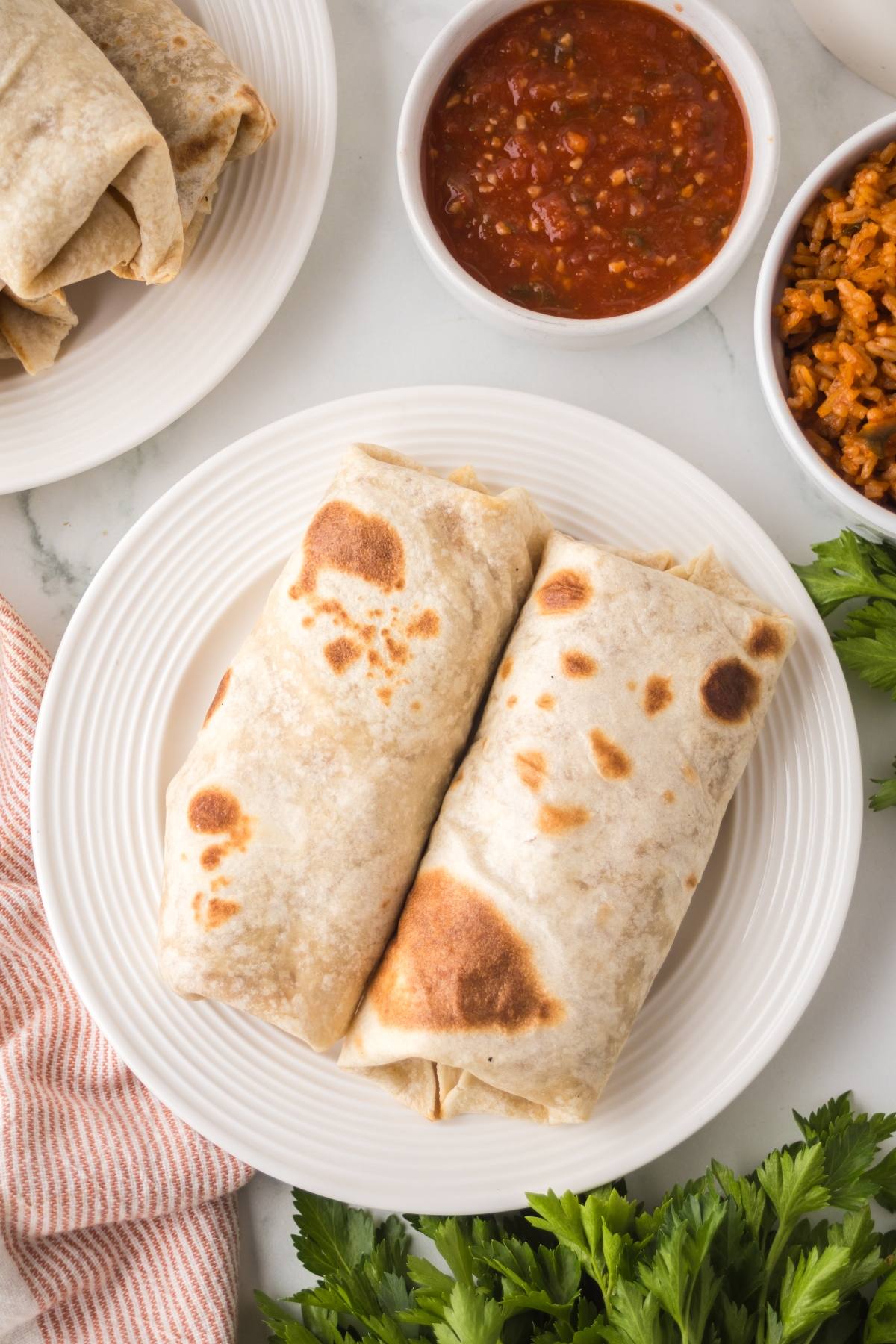 Two bean and cheese burritos on a white plate with salsa, rice, and fresh herbs nearby on a white table.