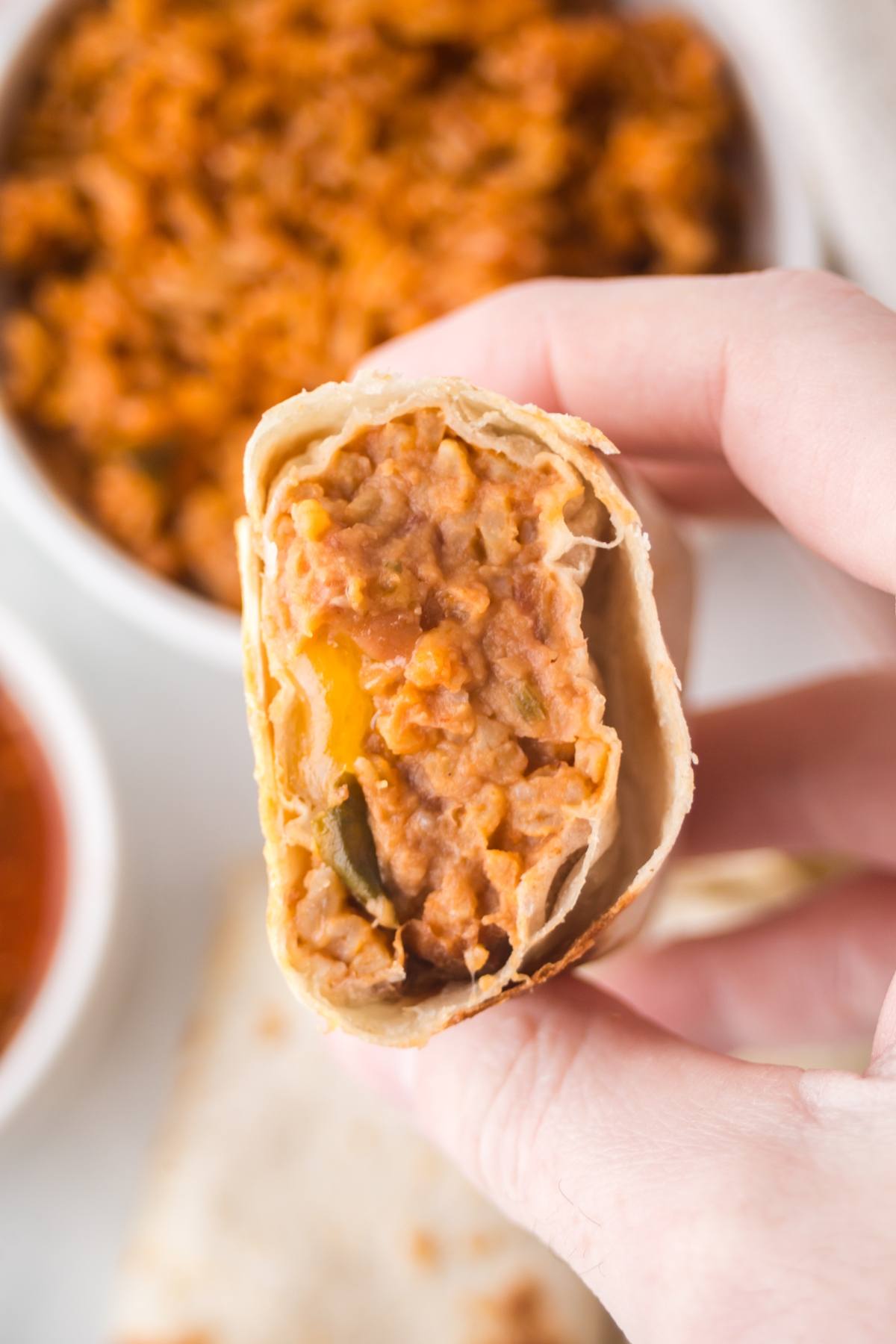 A hand holds a burrito with a visible filling of refried beans and melted cheese.