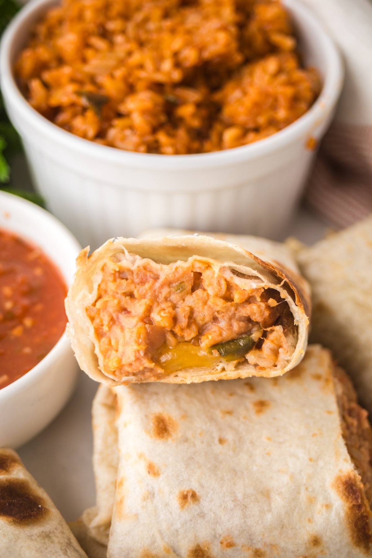 A cut bean and cheese burrito filled with rice, beans, and cheese on a plate with salsa and Mexican rice in the background.