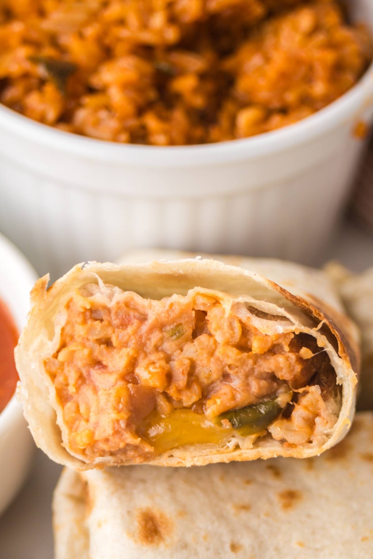 A close-up of a burrito cut open, showing a cheesy rice filling, with a bowl of rice in the background.