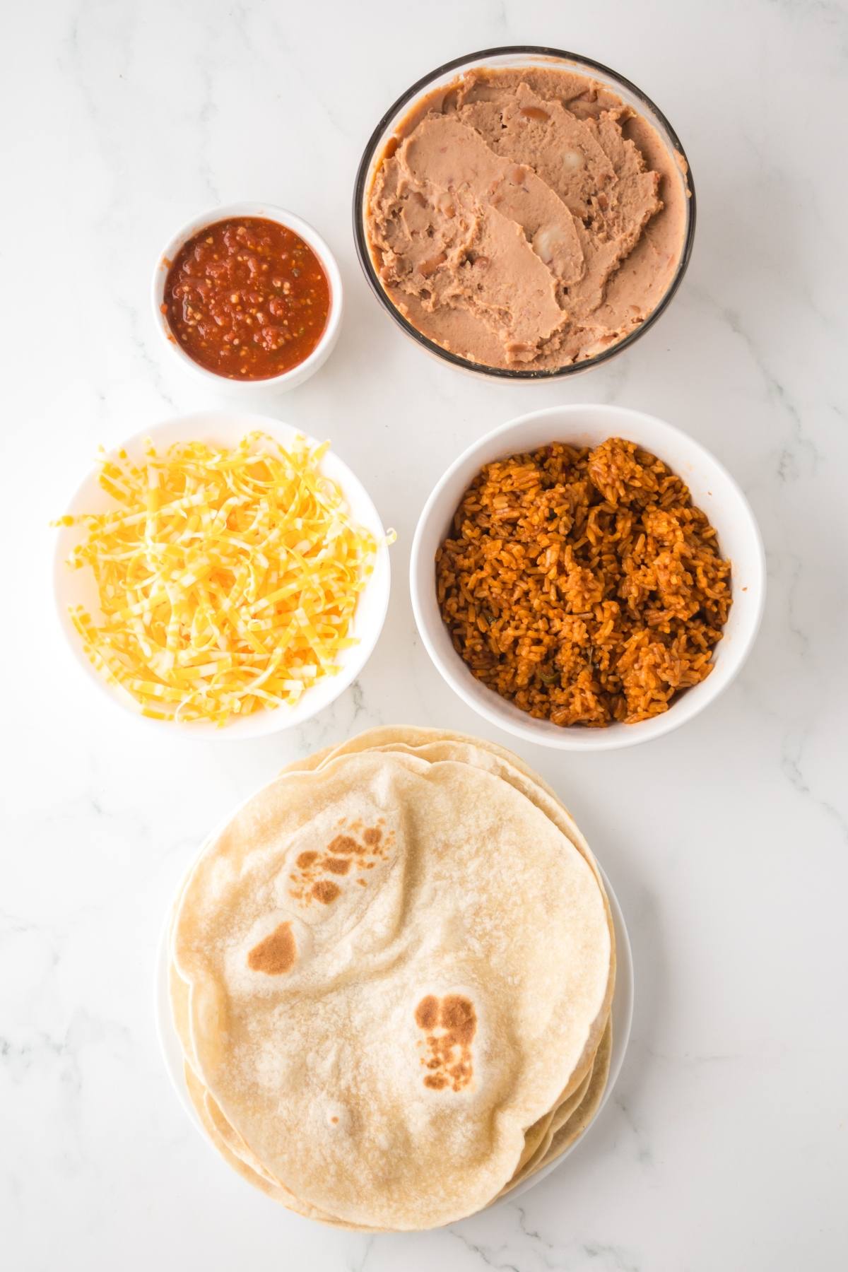 Tortillas, shredded cheese, salsa, refried beans, and rice in bowls on a white surface.