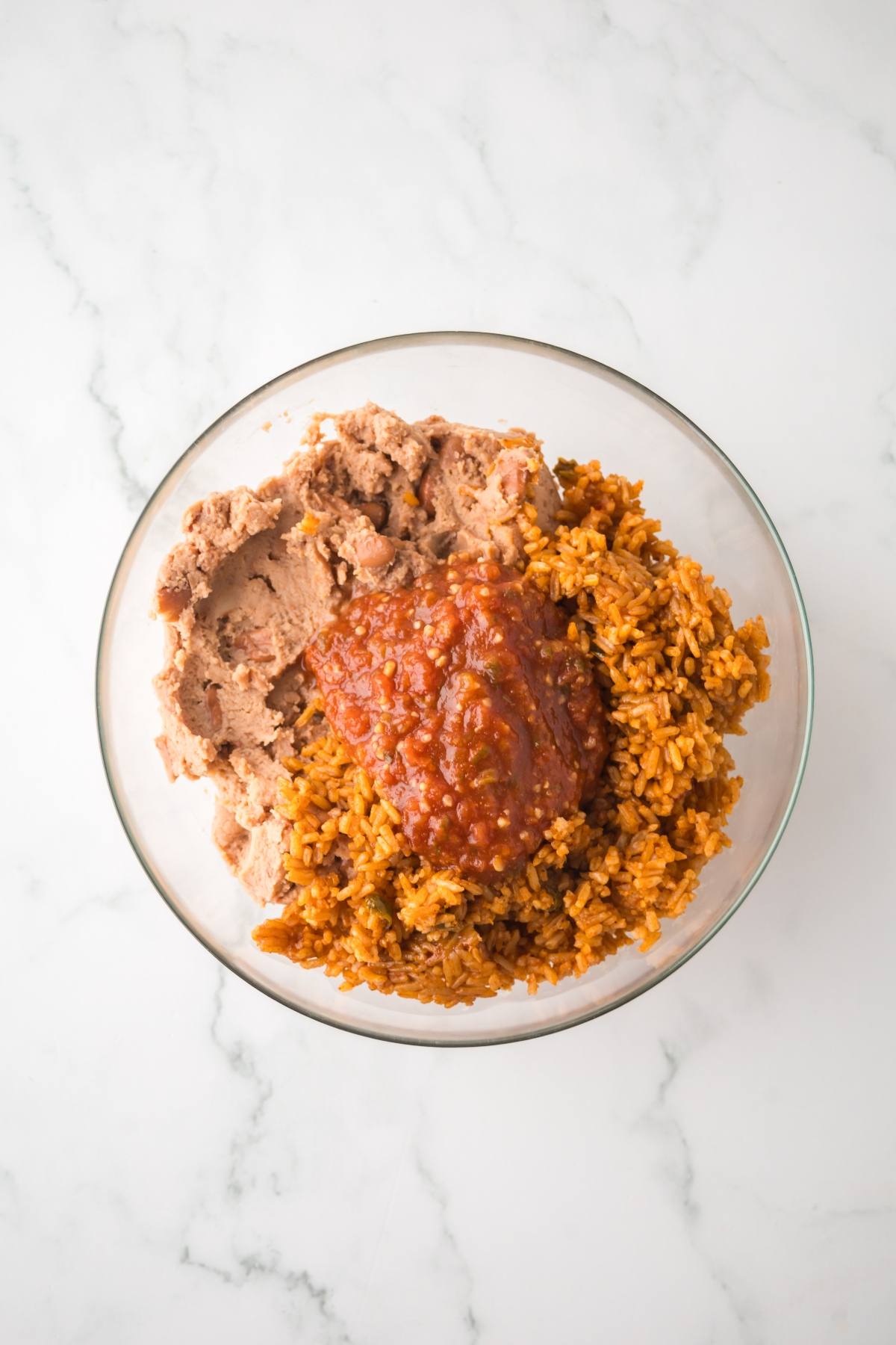 A glass bowl with refried beans, Mexican rice, and salsa on a white marble surface.