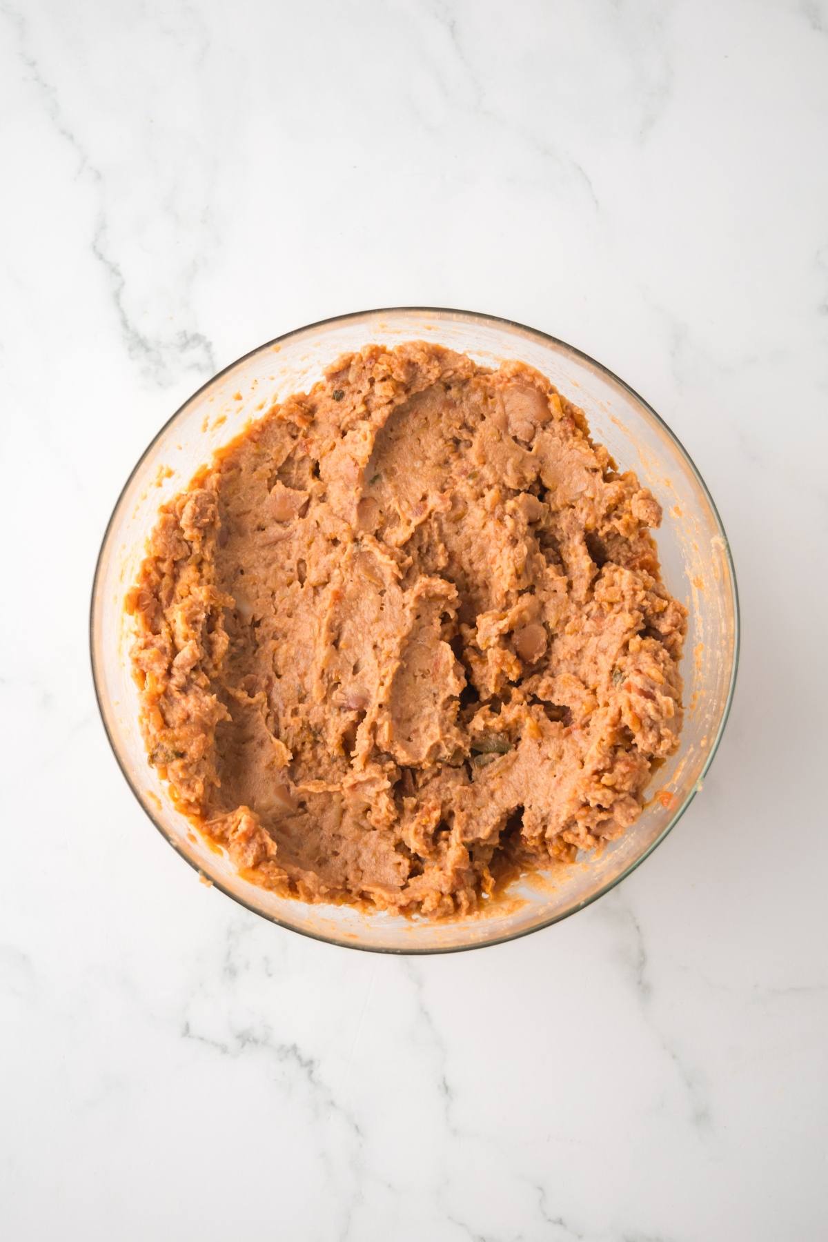 A glass bowl filled with mashed refried beans on a white marble surface.