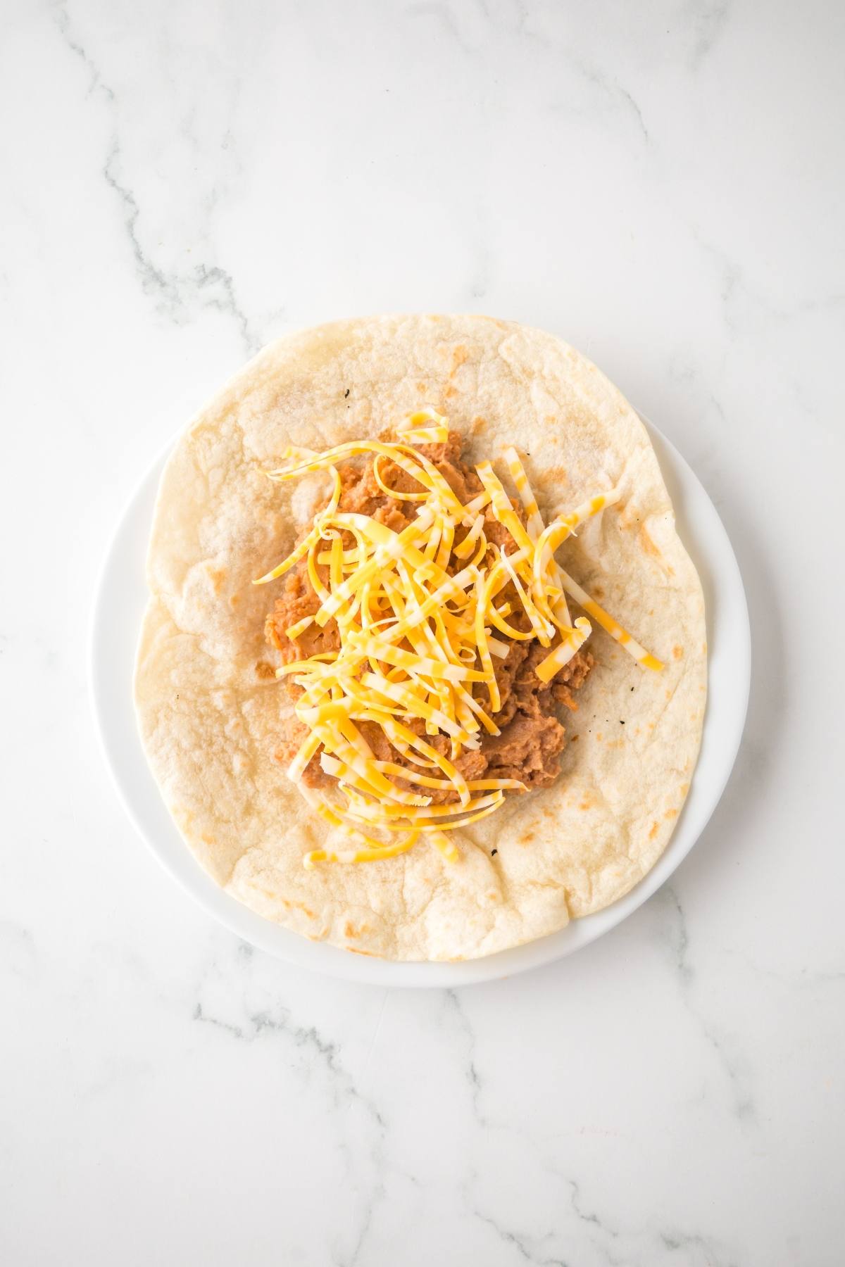 A flour tortilla with refried beans and shredded cheese on a white plate.