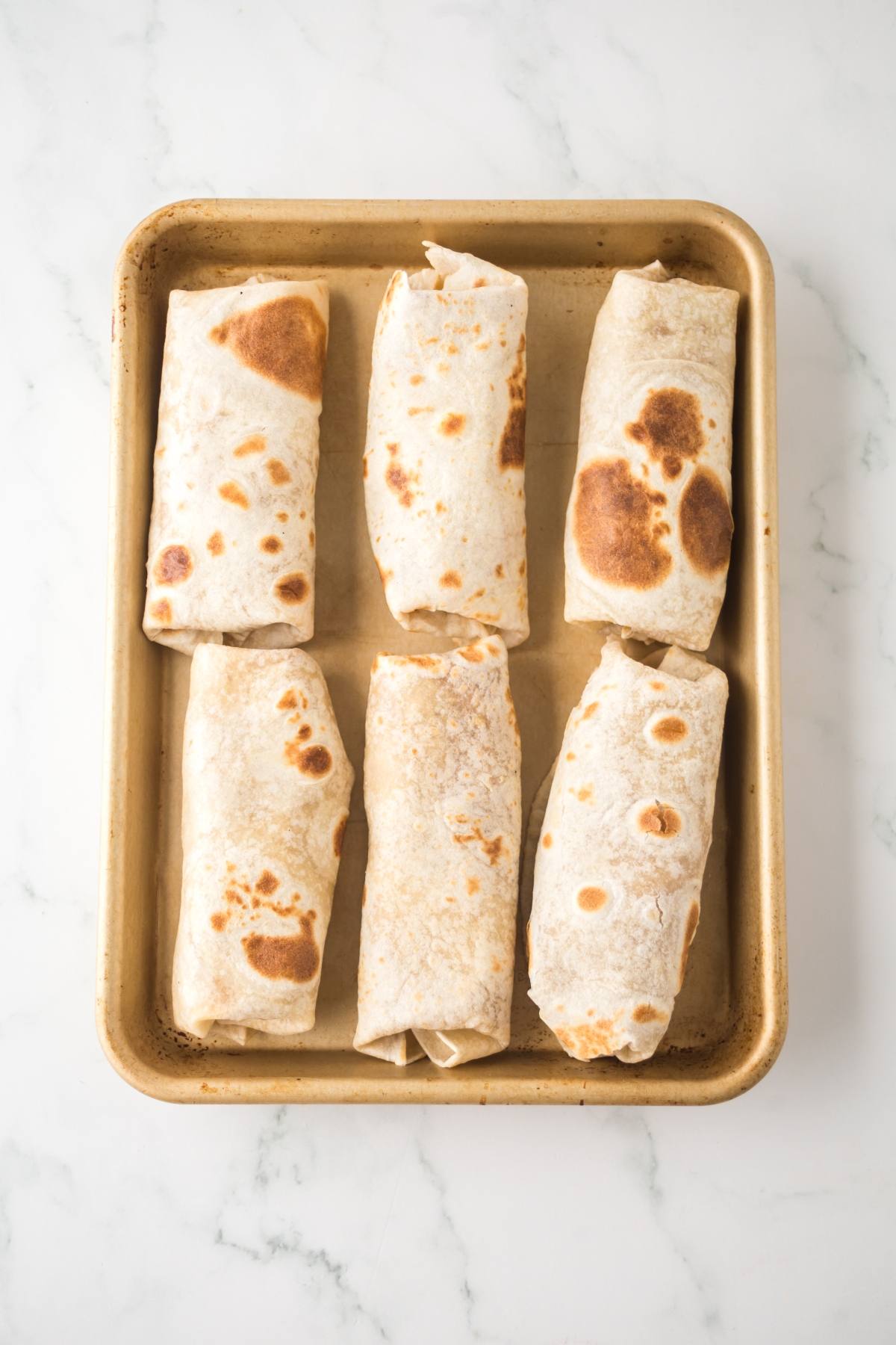 A baking tray with six rolled and toasted burritos on a white marble surface.