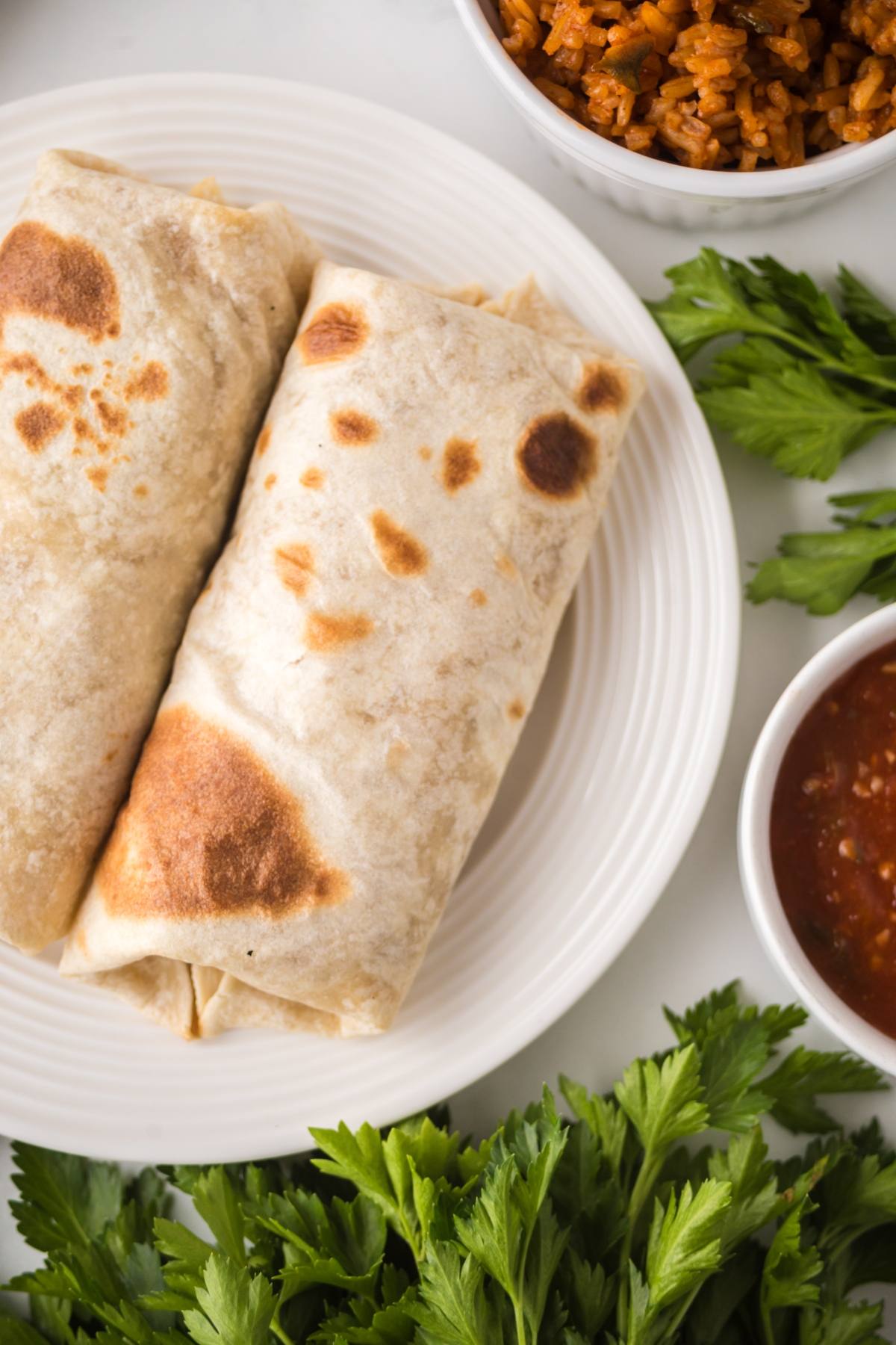 Two bean and cheese burritos on a white plate with salsa, rice, and fresh parsley on the side.