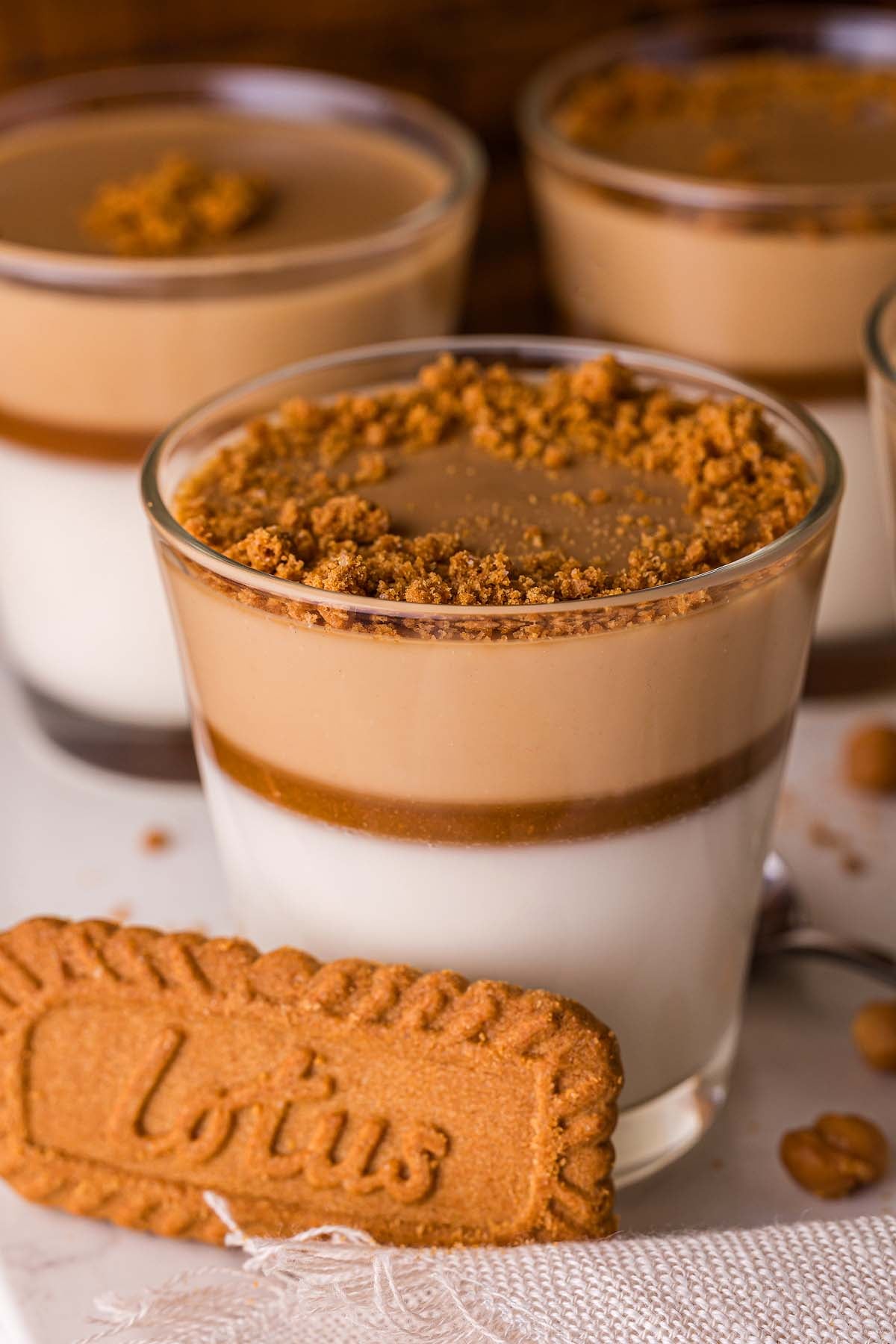 Glass of layered Biscoff Panna Cotta topped with cookie crumbs, featuring a Lotus biscuit in front and more tempting desserts in the background.