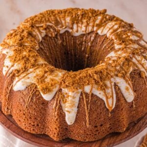 A Biscoff Cake with white icing and crumbled cookie topping sits on a wooden cake stand, surrounded by plates and cookies nearby.