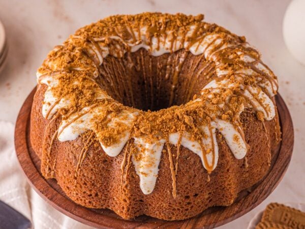 A Biscoff Cake with white icing and crumbled cookie topping sits on a wooden cake stand, surrounded by plates and cookies nearby.