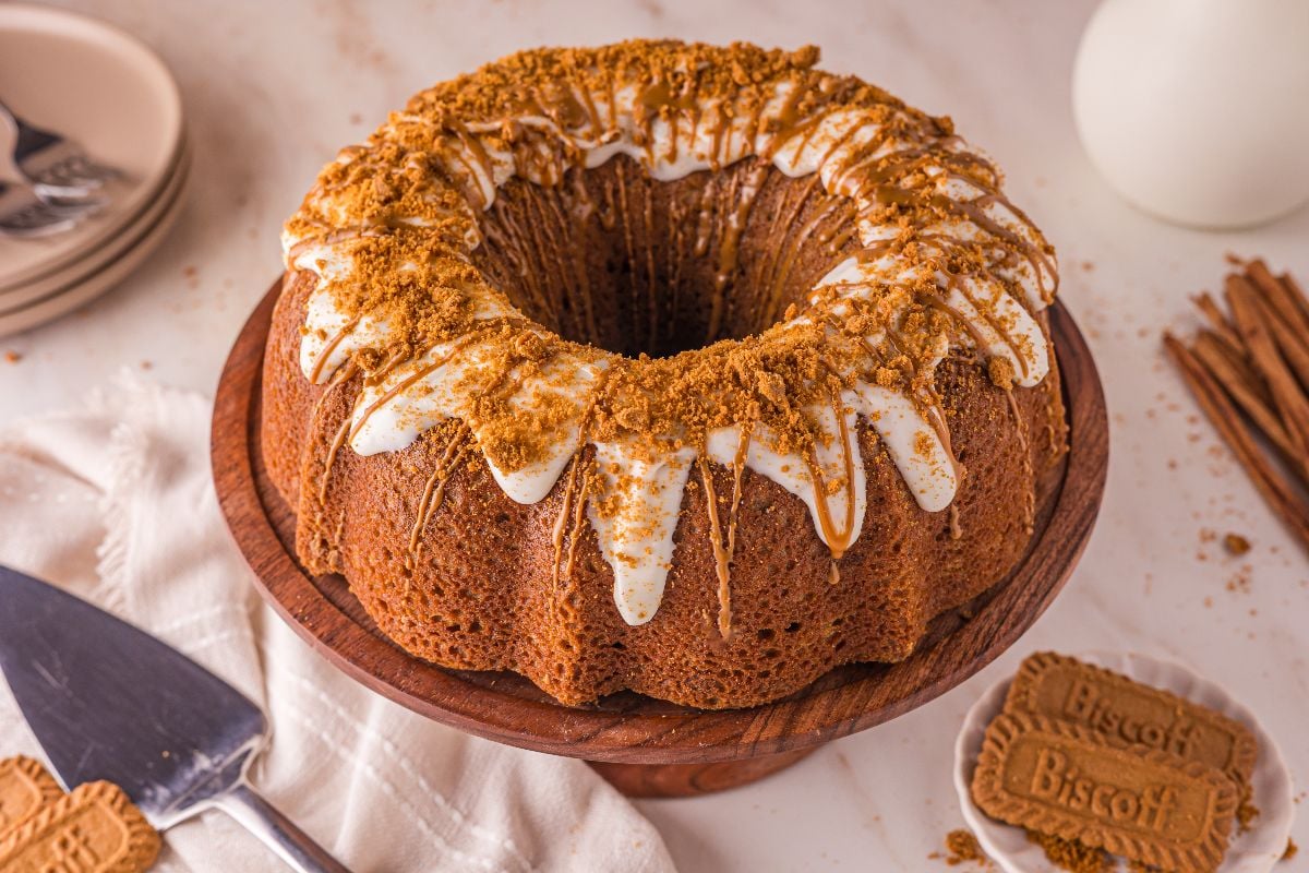A Biscoff Cake Bundt with white icing and crushed cookies on top, displayed on a wooden stand with Biscoff cookies nearby.