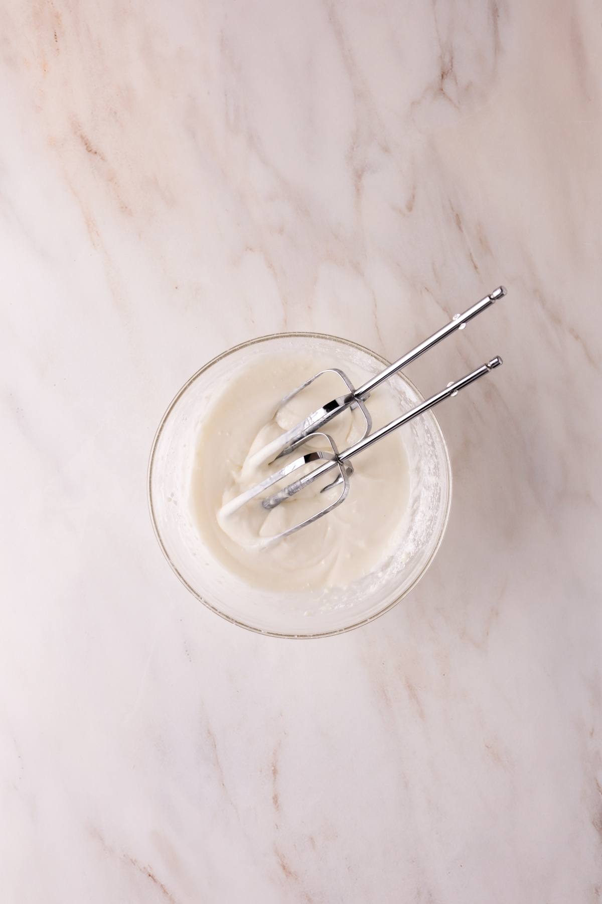 A glass bowl with whipped cream and two electric mixer beaters on a marble countertop.
