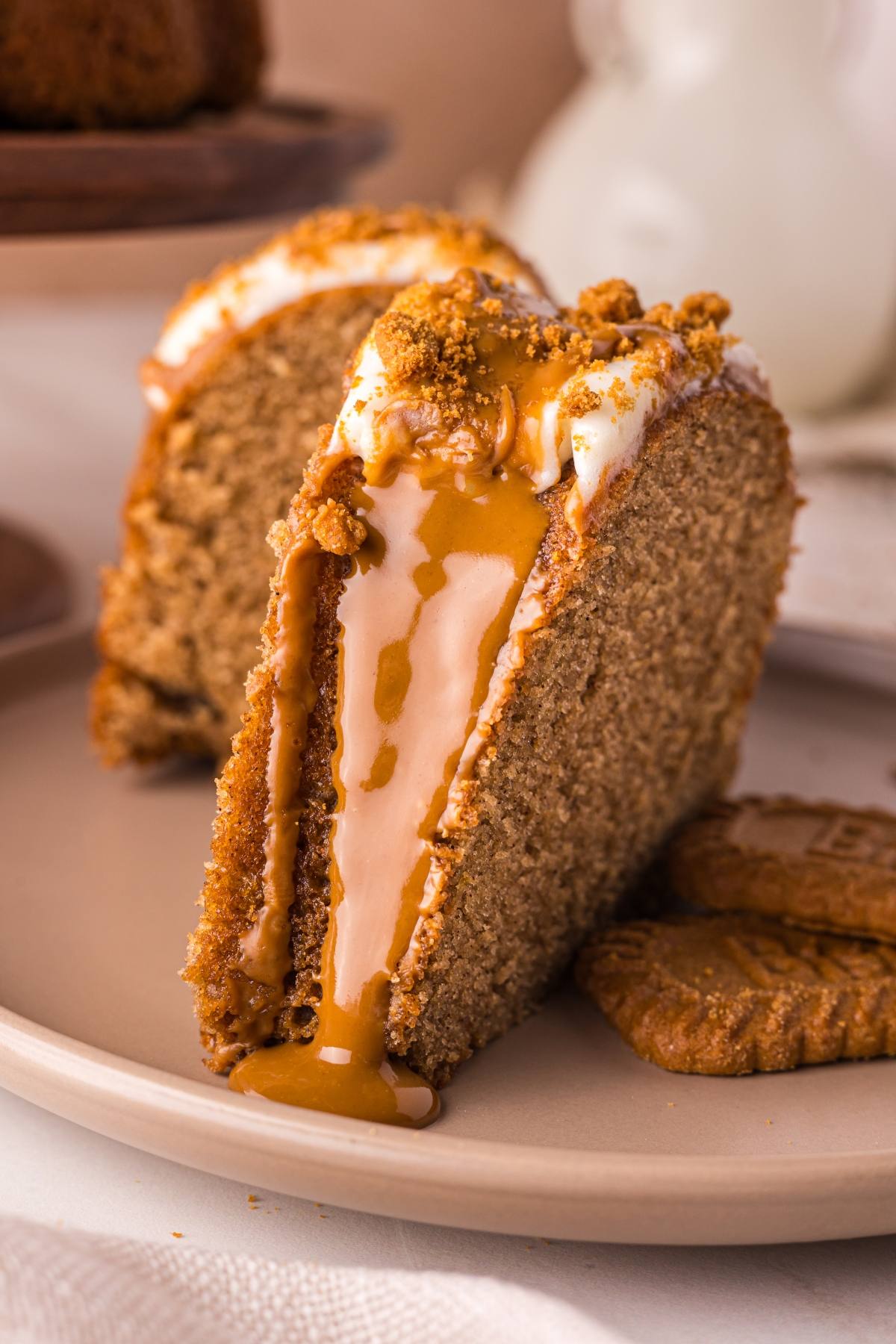 A slice of spiced biscoff cake with creamy glaze and cookie crumbs, served on a plate with cookies beside it.