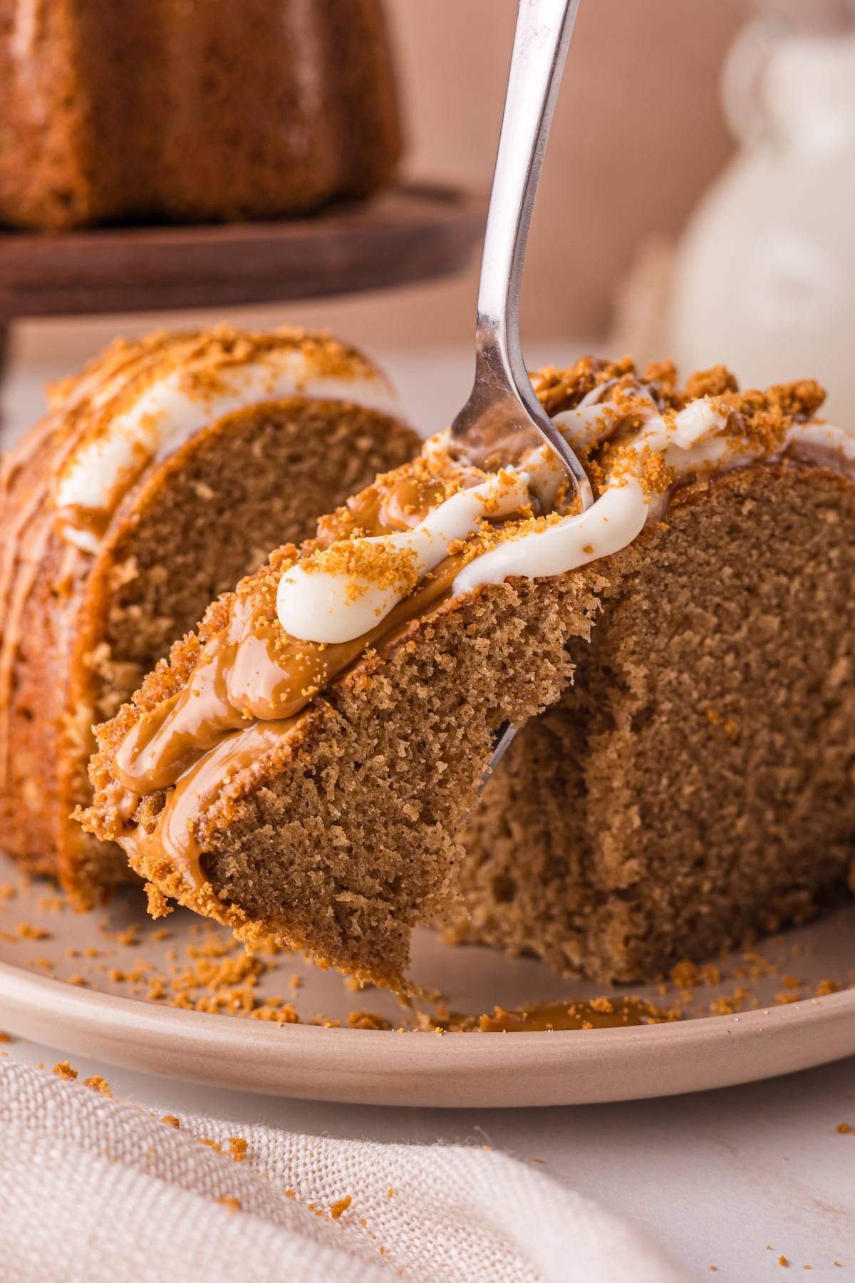A fork lifts a slice of spiced biscoff cake topped with white and caramel icing from a plate.