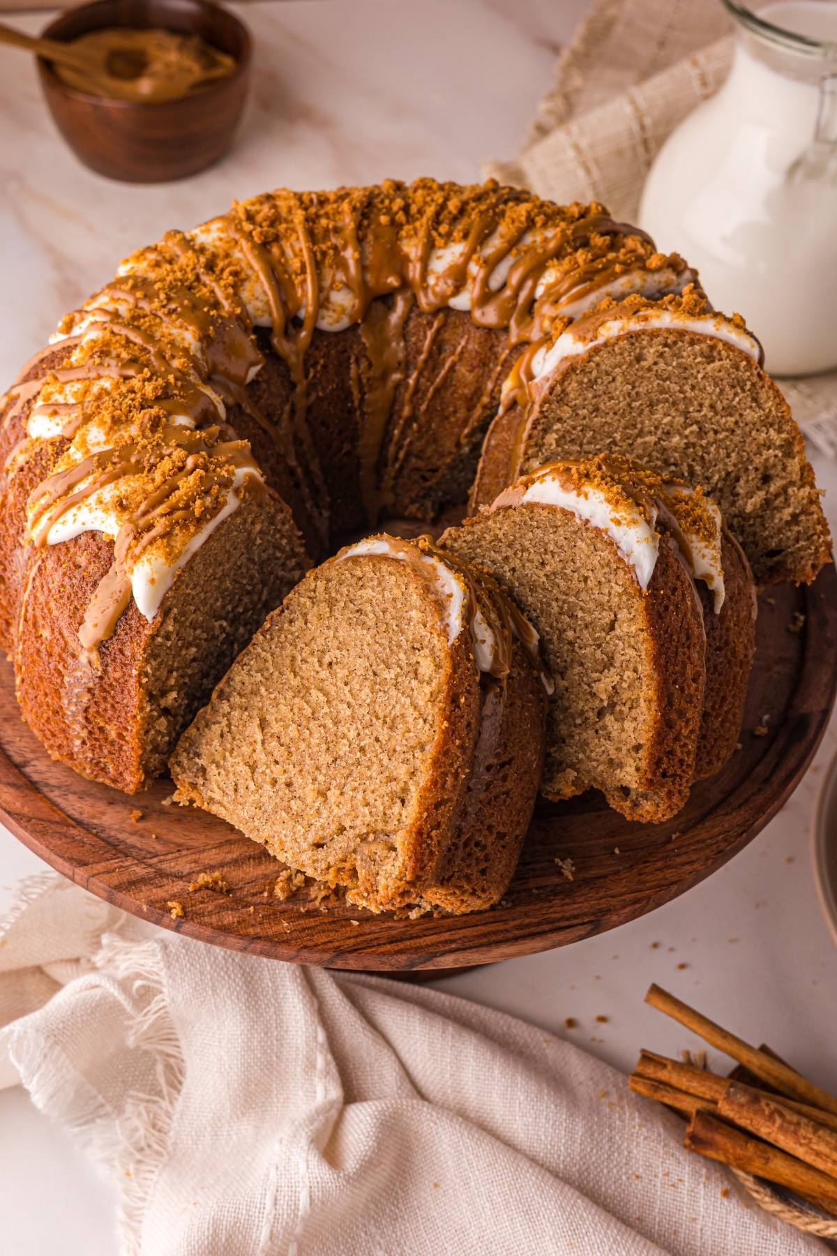 biscoff cake with icing and crumbs on top, partially sliced on a wooden cake stand, with milk and cinnamon sticks nearby.