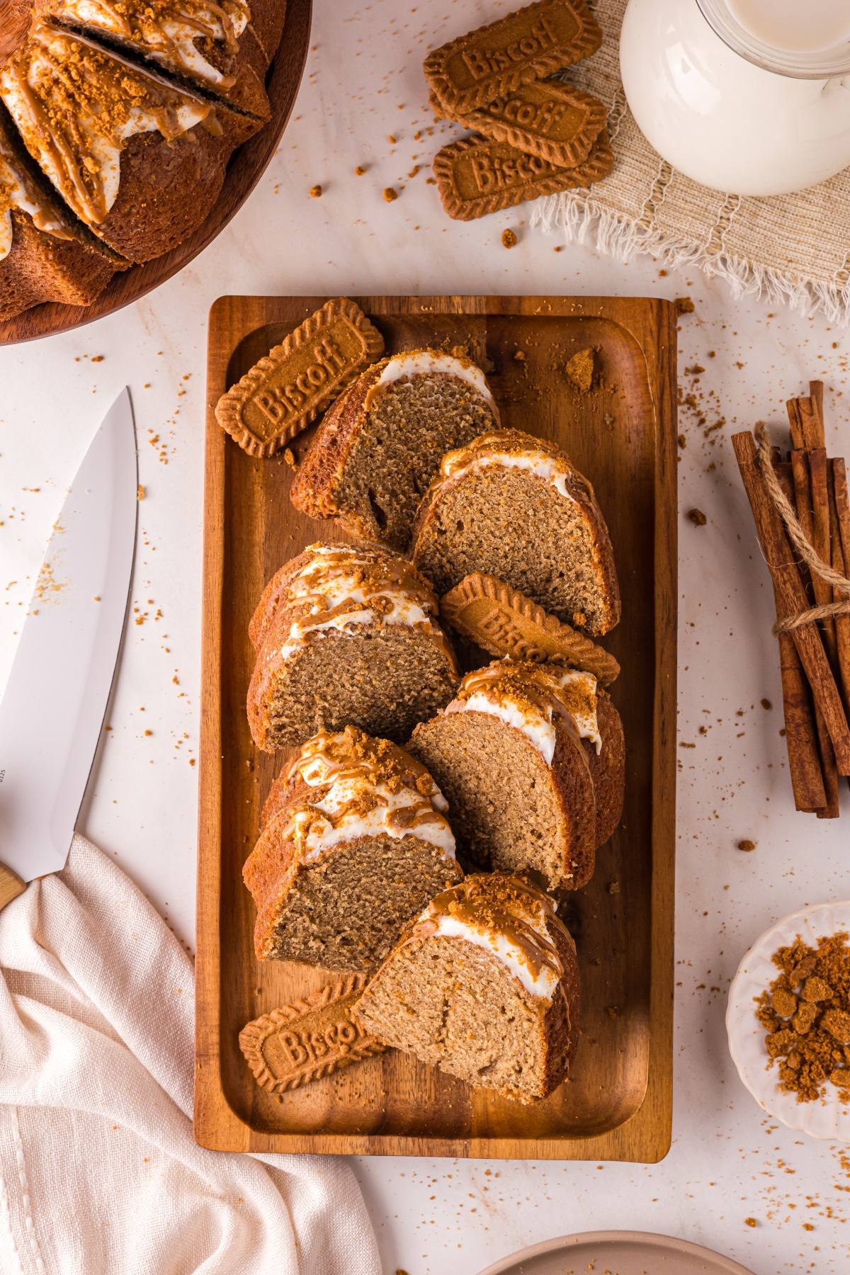 Sliced biscoff cake with icing and crushed cookies on a wooden tray, surrounded by Biscoff cookies and cinnamon sticks.