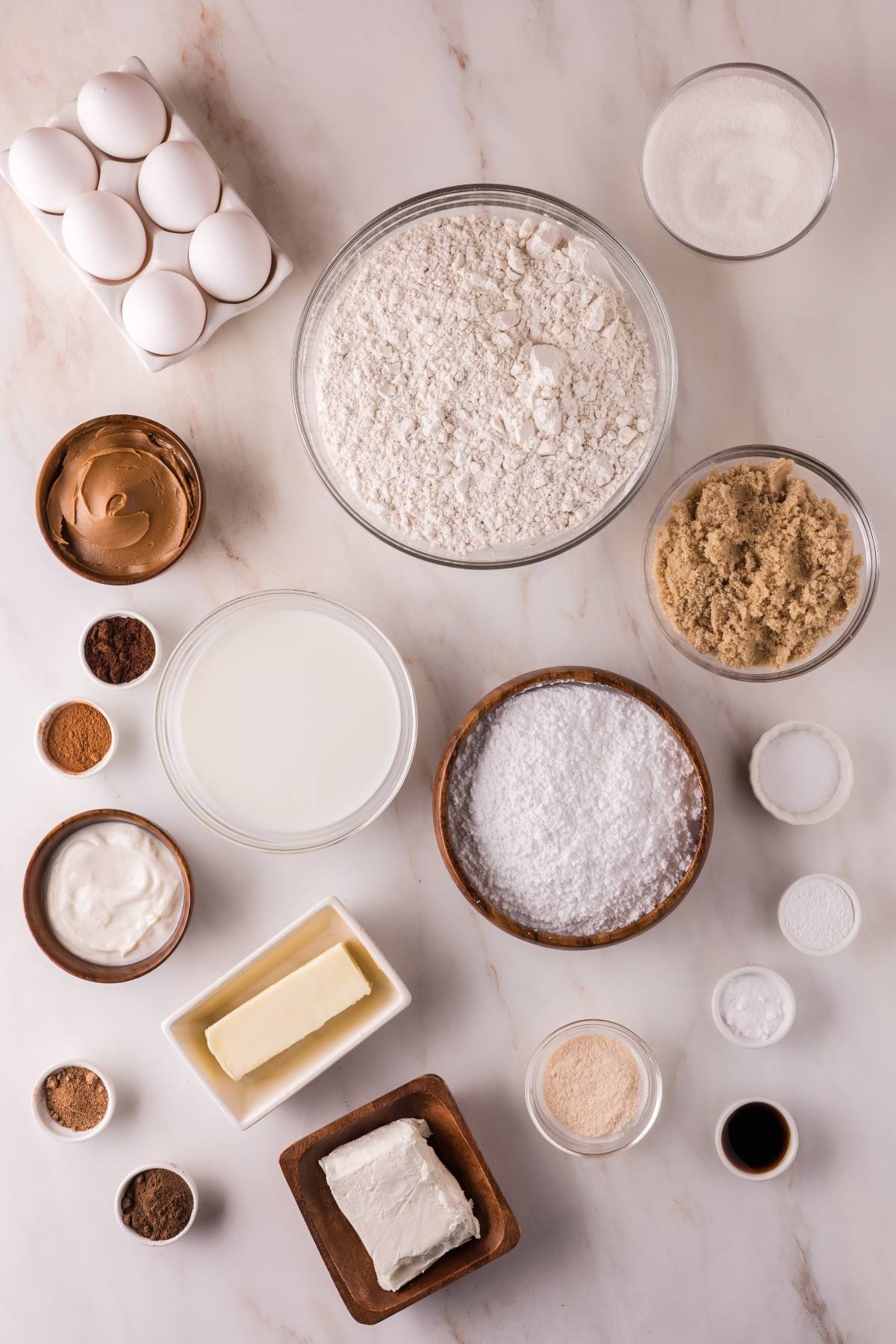 Baking ingredients on a marble surface: eggs, flour, sugars, butter, cream cheese, spices, milk, and vanilla.