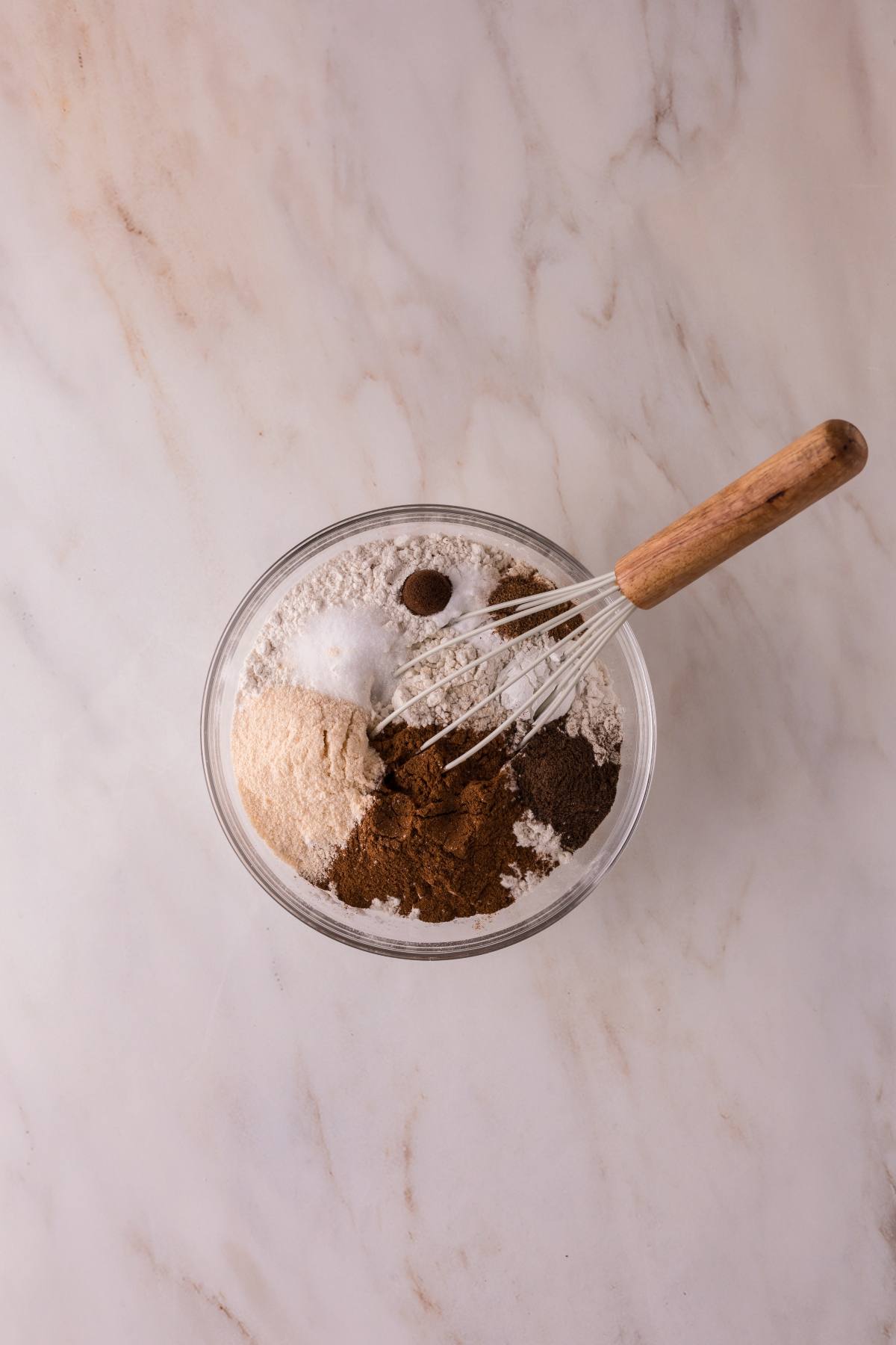 A glass bowl with dry baking ingredients and a whisk on a marble surface.