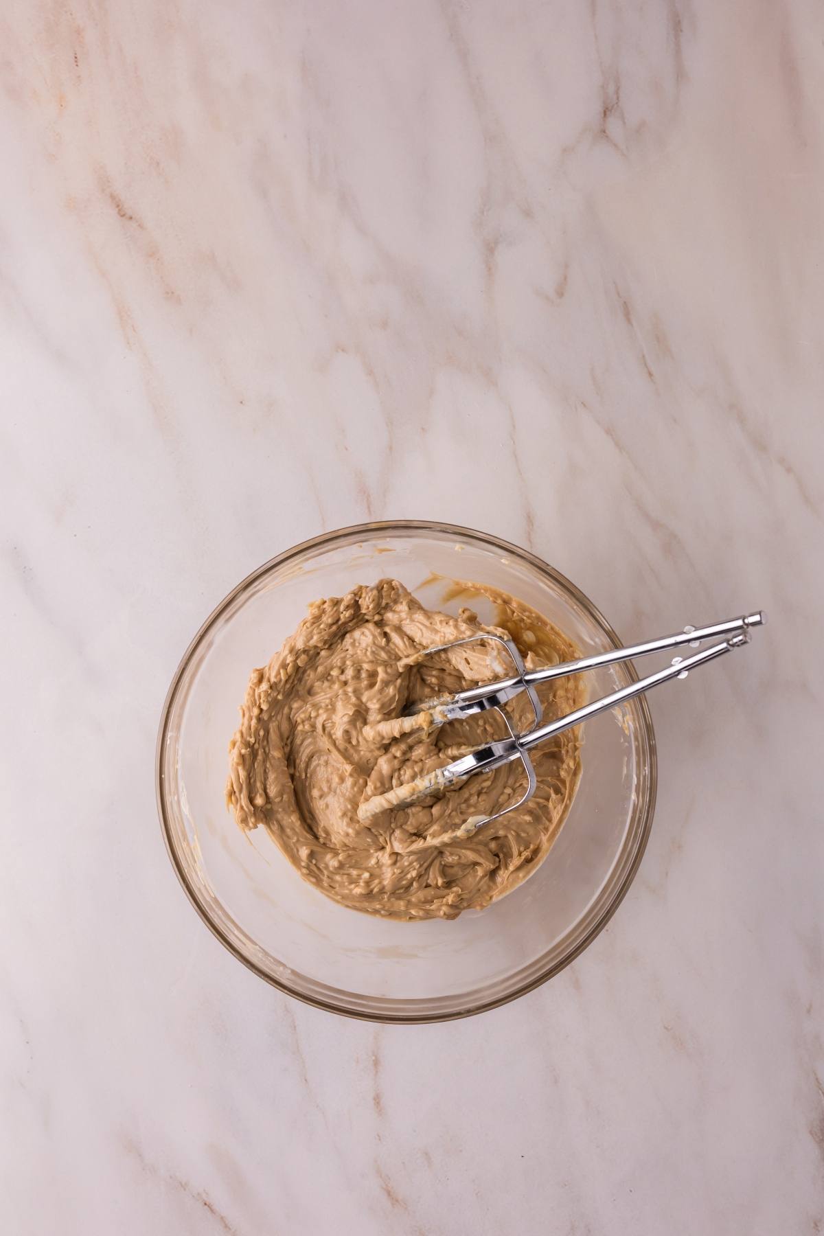 A glass bowl with creamy batter and a metal hand mixer on a light marble surface.