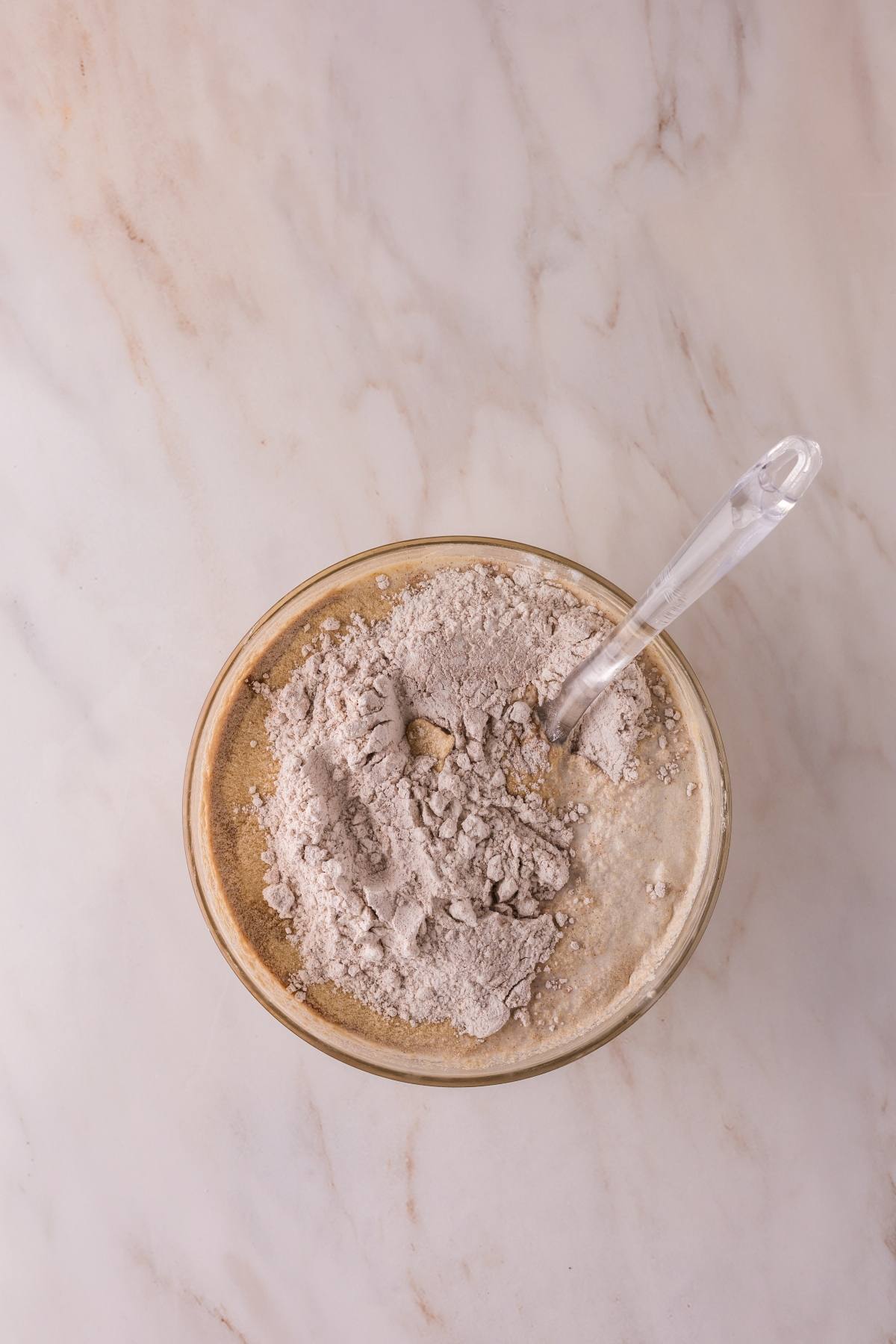 A glass mixing bowl with flour and batter, and a clear spoon, on a marble countertop.