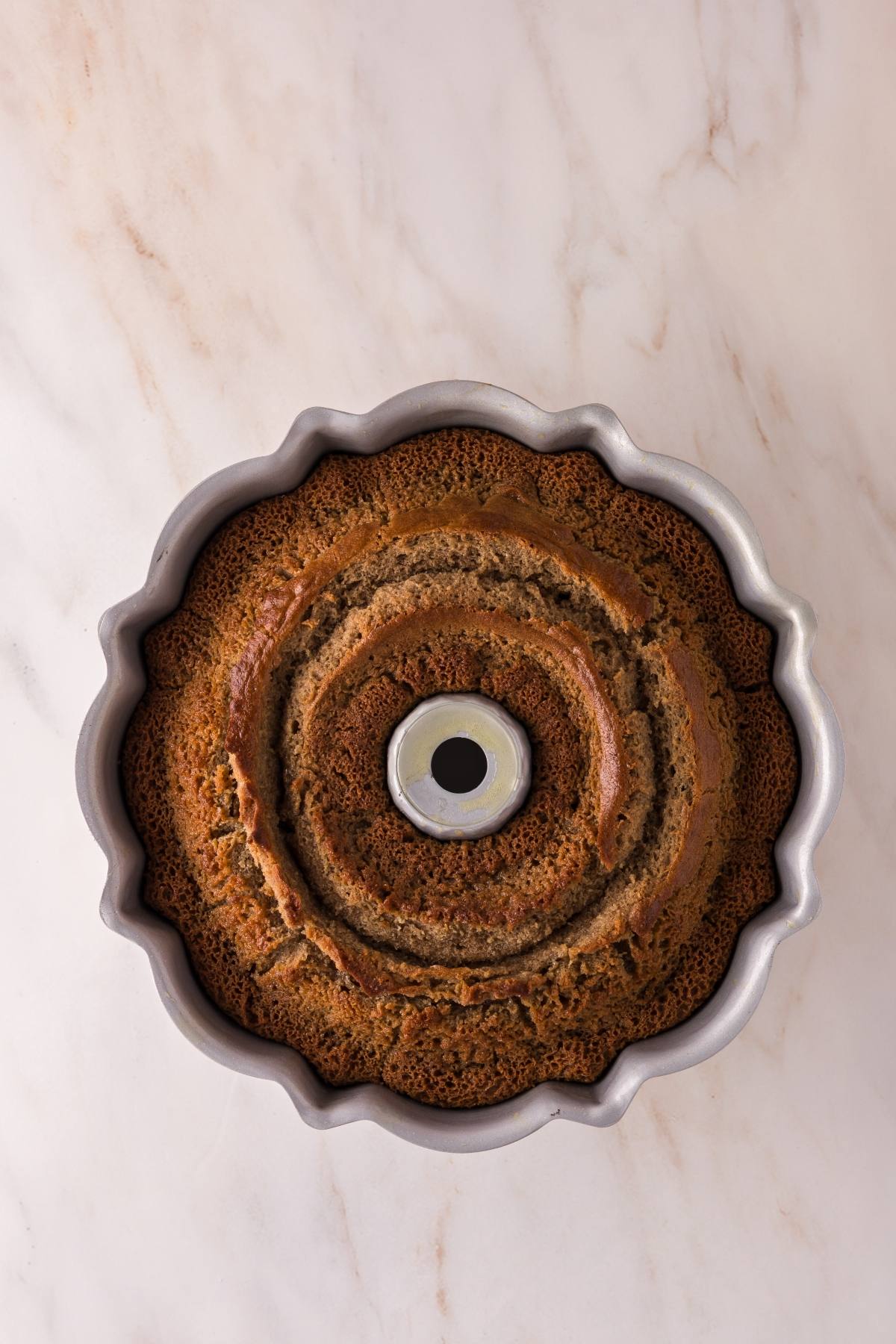 A baked bundt cake in a silver pan on a marble surface, viewed from above.