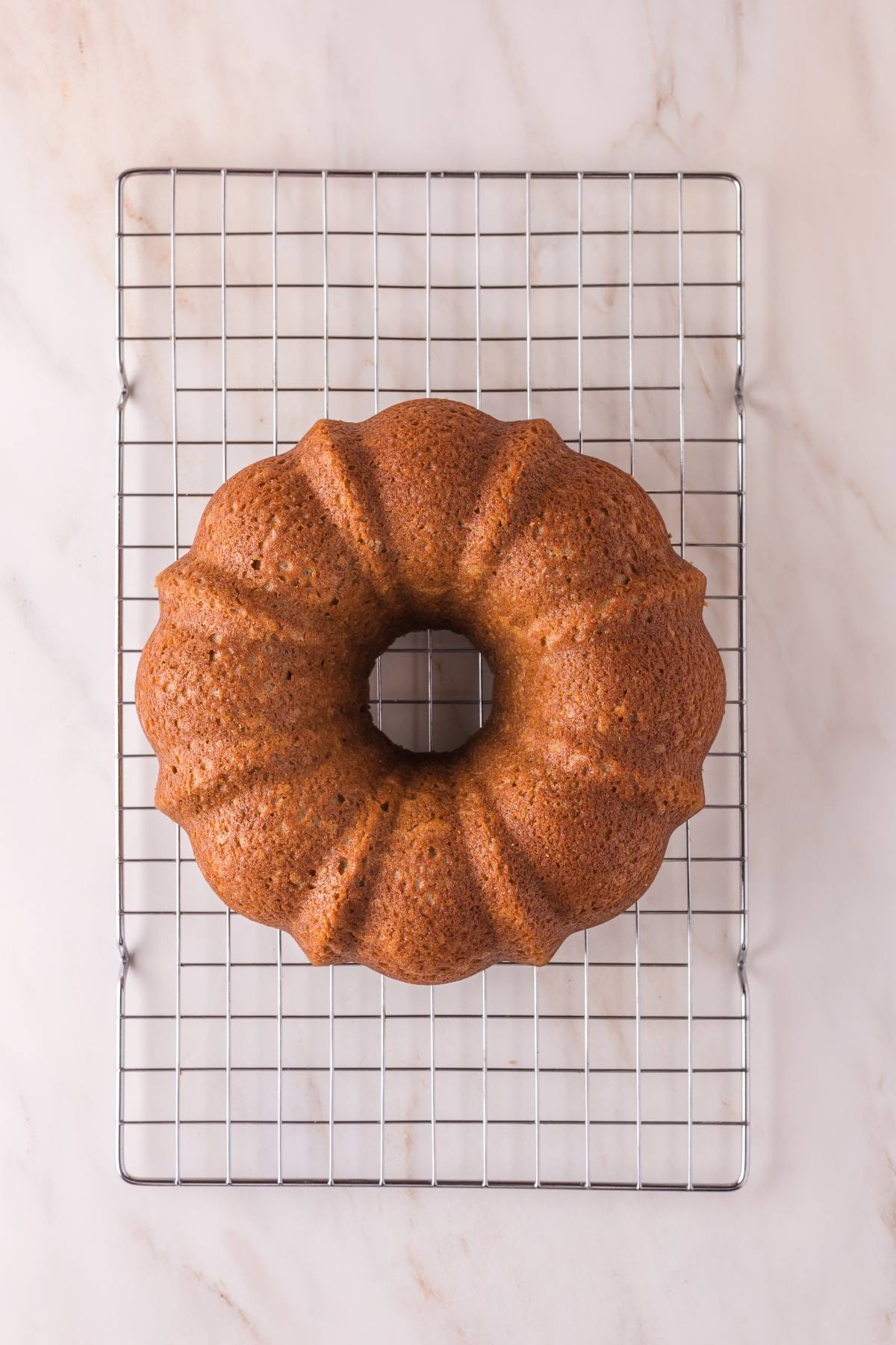 A golden brown bundt cake cooling on a metal rack over a light marble surface.