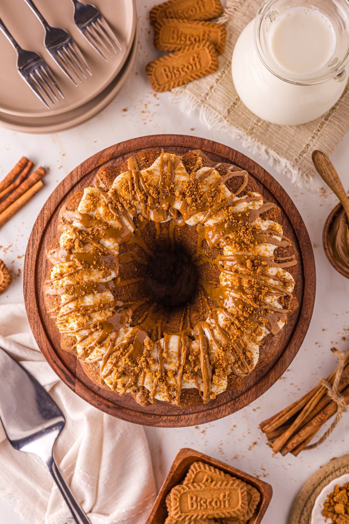 A Bundt cake topped with icing and biscuit crumbs, surrounded by cookies, cinnamon sticks, and a jug of milk.
